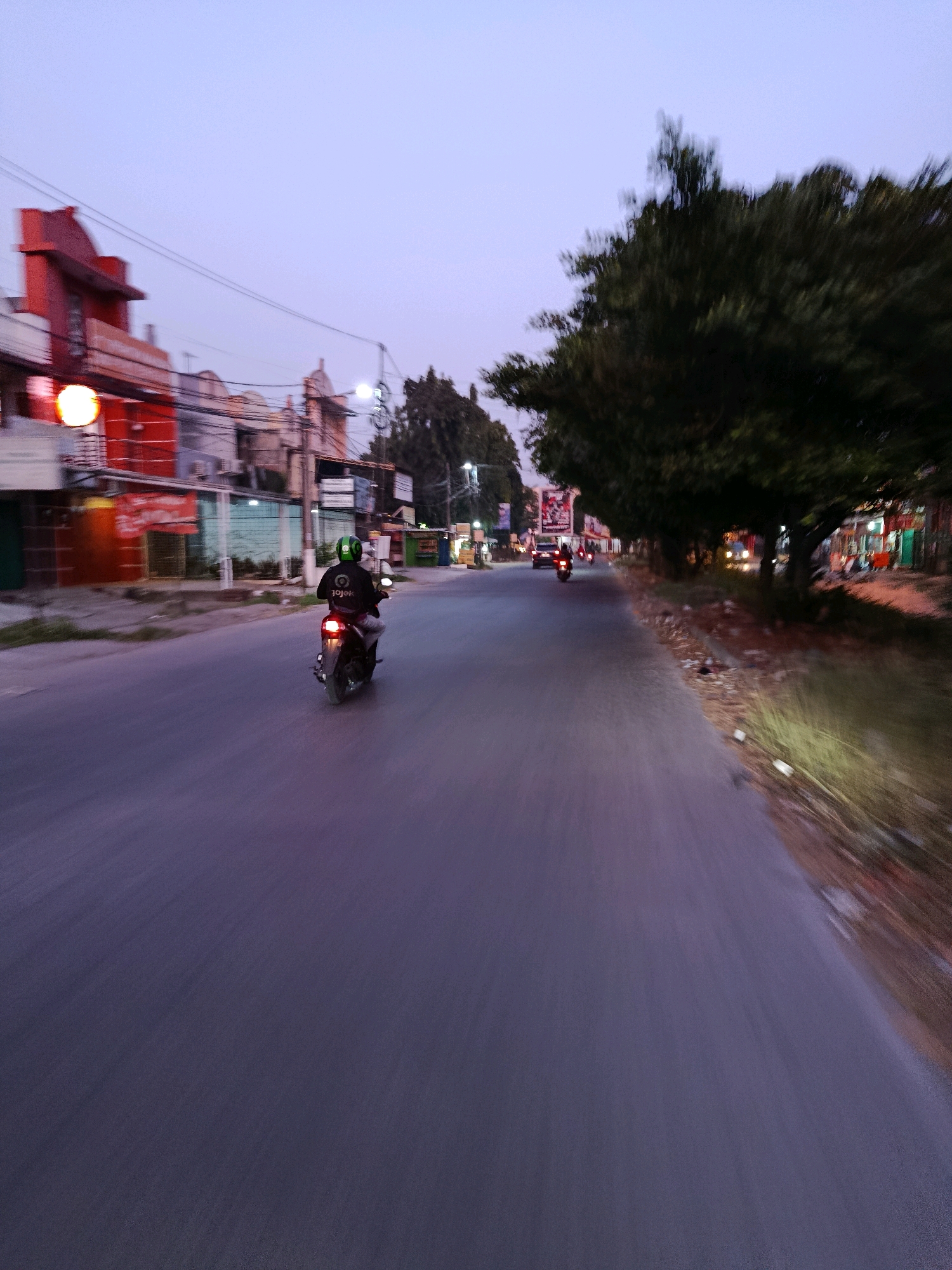 A motorcycle rider wearing a green helmet and a black jacket with the "Gojek" logo is traveling on a two-lane road in Bekasi, Indonesia. The rider is in the right lane, moving away from the camera. The surrounding environment appears to be urban with buildings on the left side of the road, including a red building with illuminated signs. The right side of the road features a sidewalk lined with trees and more buildings, some of which have lit windows. The sky is a dusky purple, suggesting it is either early morning or late evening. Other vehicles, including another motorcycle and a car, are visible further down the road. The motion blur indicates the photo was taken from a moving vehicle.