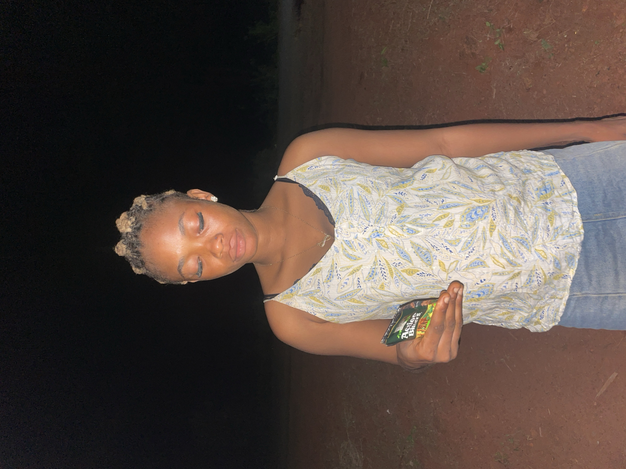 A young woman stands in a dimly lit area near Ogbe, Nigeria. She is facing forward and slightly angled to her left. She has blonde hair styled in small twists, and she is wearing a light-colored, patterned tank top with thin straps, blue jeans, and a delicate gold necklace. Her eyes are closed. She holds a packet of "Action Bitters" in her right hand. The background is dark, with some greenery visible behind her. The ground appears to be red dirt.