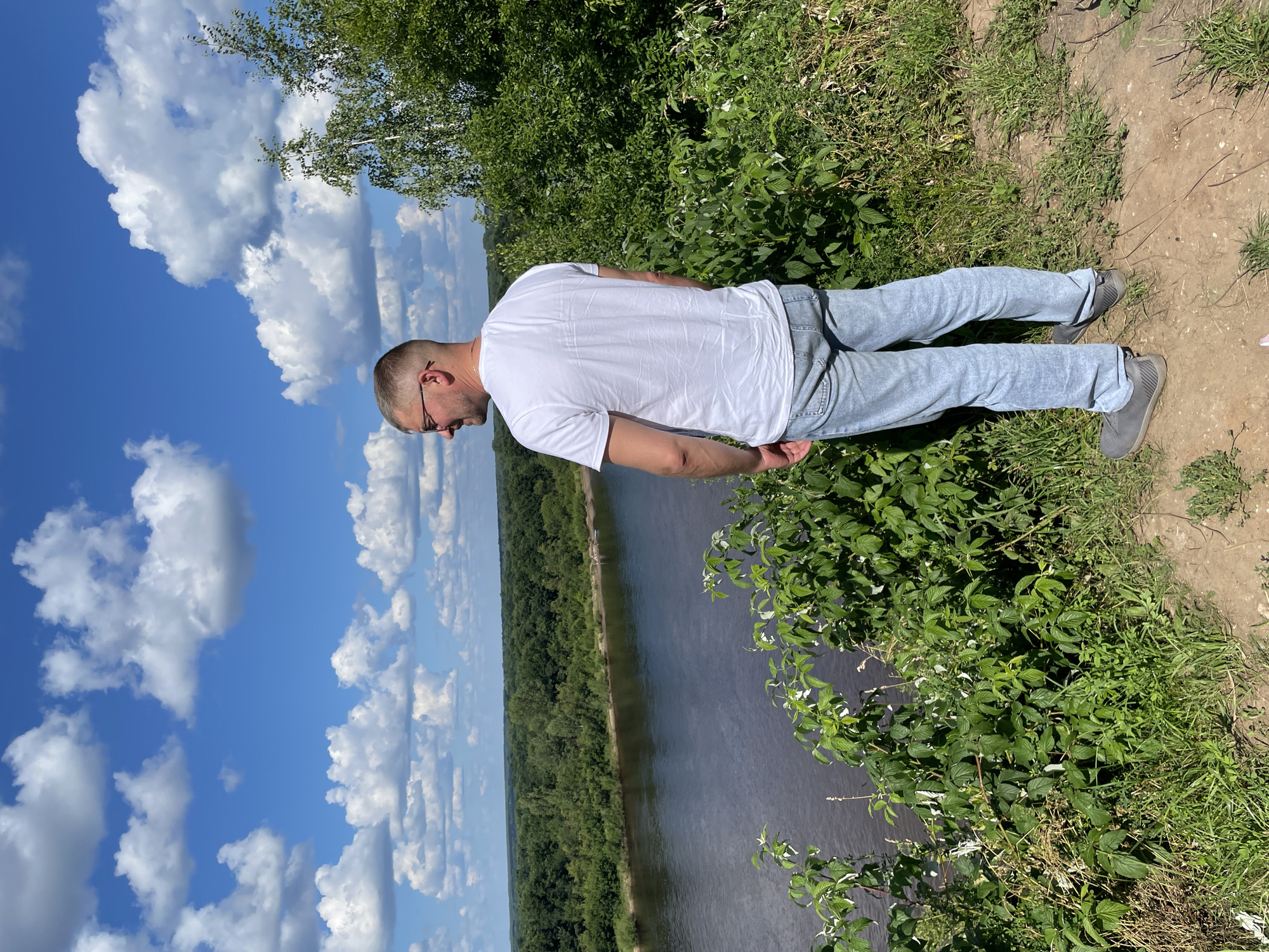 A man with short blonde hair and glasses stands on a grassy hill overlooking a wide river. He is wearing a white t-shirt and light blue jeans, with his back to the camera. The river is dark and reflects the blue sky, which is dotted with fluffy white clouds. Lush green trees and vegetation line the riverbank and the hill where the man is standing. The background shows a dense forest stretching to the horizon. The scene appears to be a natural landscape with no visible buildings or urban elements, suggesting a location outside of the city center of Kirovo-Chepetsk, Russia, possibly in a scenic natural park or rural area.