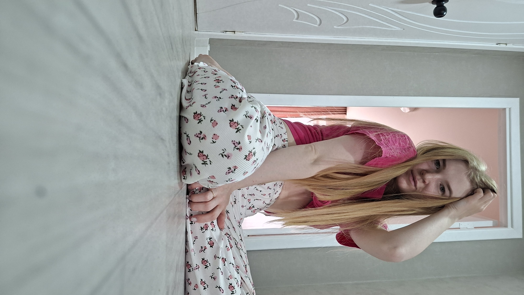 The image, originally oriented on its side, captures a young woman posing in a relaxed, seated position on a light-colored floor within an indoor setting, likely in Chaikovsky, Russia. She has long, straight blonde hair and light-colored eyes, looking directly at the camera with a calm and subtle smile. She is wearing a bright pink top and white floral patterned pants featuring small pink roses and green leaves. One of her hands rests on the floor beside her, while the other is gently touching her hair near her head.
The background shows a white doorway with decorative molding and a black doorknob. Through this doorway, an adjacent room with light pink walls is visible. The scene is illuminated by diffused light, suggesting it is daytime indoors. No text is visible in the image. The overall impression is a casual portrait taken in a domestic environment.