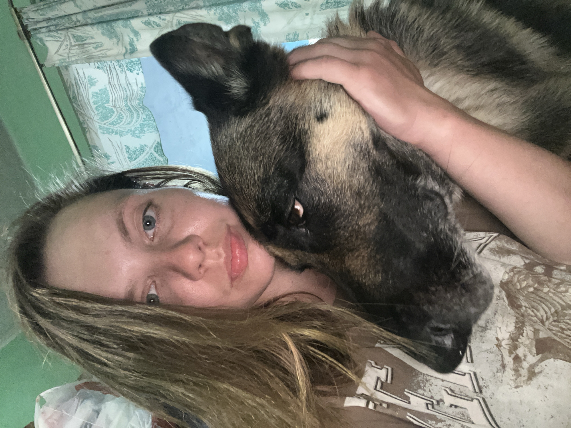 A close-up selfie shows a woman and a large dog in an intimate embrace, conveying warmth and affection. The woman, with fair skin, light blue eyes, and long brown hair with blonde highlights, lies on her back, looking directly at the camera with a soft smile. Her lips are a light pink.
Beside and partially on top of her, a large dog, resembling a German Shepherd with mottled dark brown, black, and tan fur, rests its head close to her face and chest. One of the dog's eyes, a deep brown, is visible, looking towards the camera. The woman's arm and hand are visible, gently holding the dog around its neck or shoulder.
The setting is indoors, likely a home, during daytime, indicated by the natural light filtering through a window in the background. The window features light blue and white patterned blinds or curtains, adorned with a design depicting trees or a natural landscape. A soft green wall is visible to the left. The woman is wearing a light-colored top, possibly a t-shirt, which has a stylized, complex pattern in darker shades across the chest. Parts of what appear to be letters, possibly "E" and "Z," are discernible within this abstract design. The scene evokes a strong bond between the woman and her pet, set within what appears to be a cozy indoor environment in Nizhny Tagil, Russia.