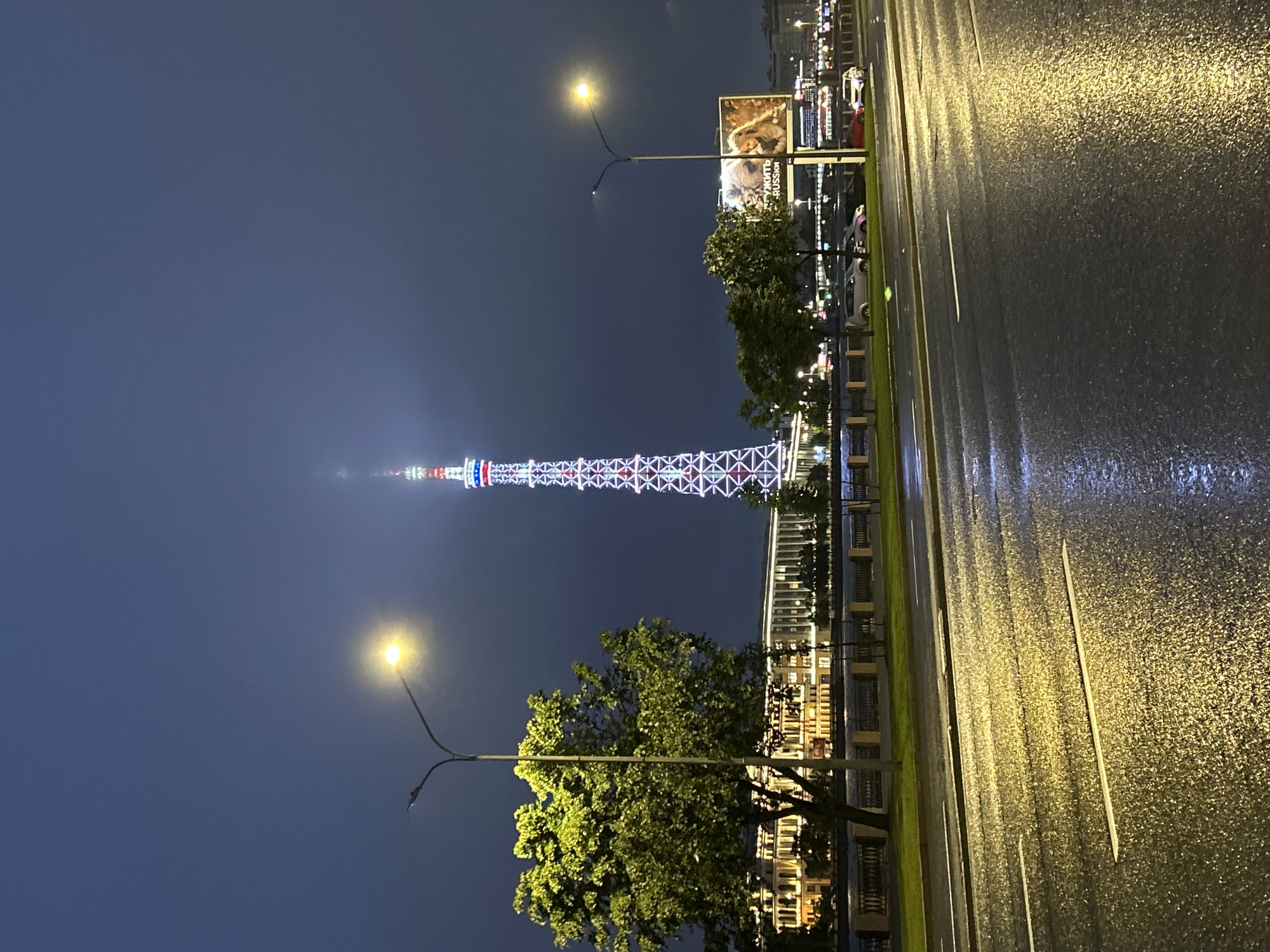 The image captures a nighttime city scene with a tall, illuminated tower as the focal point. The tower is a lattice structure with white and red lights, suggesting a significant landmark. In the foreground, a wet, reflective asphalt road is visible, indicating recent rain. Streetlights cast a yellow glow on the road and the surrounding area. To the left of the tower, a large, leafy tree is illuminated, and behind it, a long, multi-story building with lit windows runs along a body of water, likely a river or canal. A billboard with an image of a woman is positioned to the right of the tower. Several cars are parked and driving along the road and on the other side of the street. The sky is dark and cloudy, typical of a night sky.