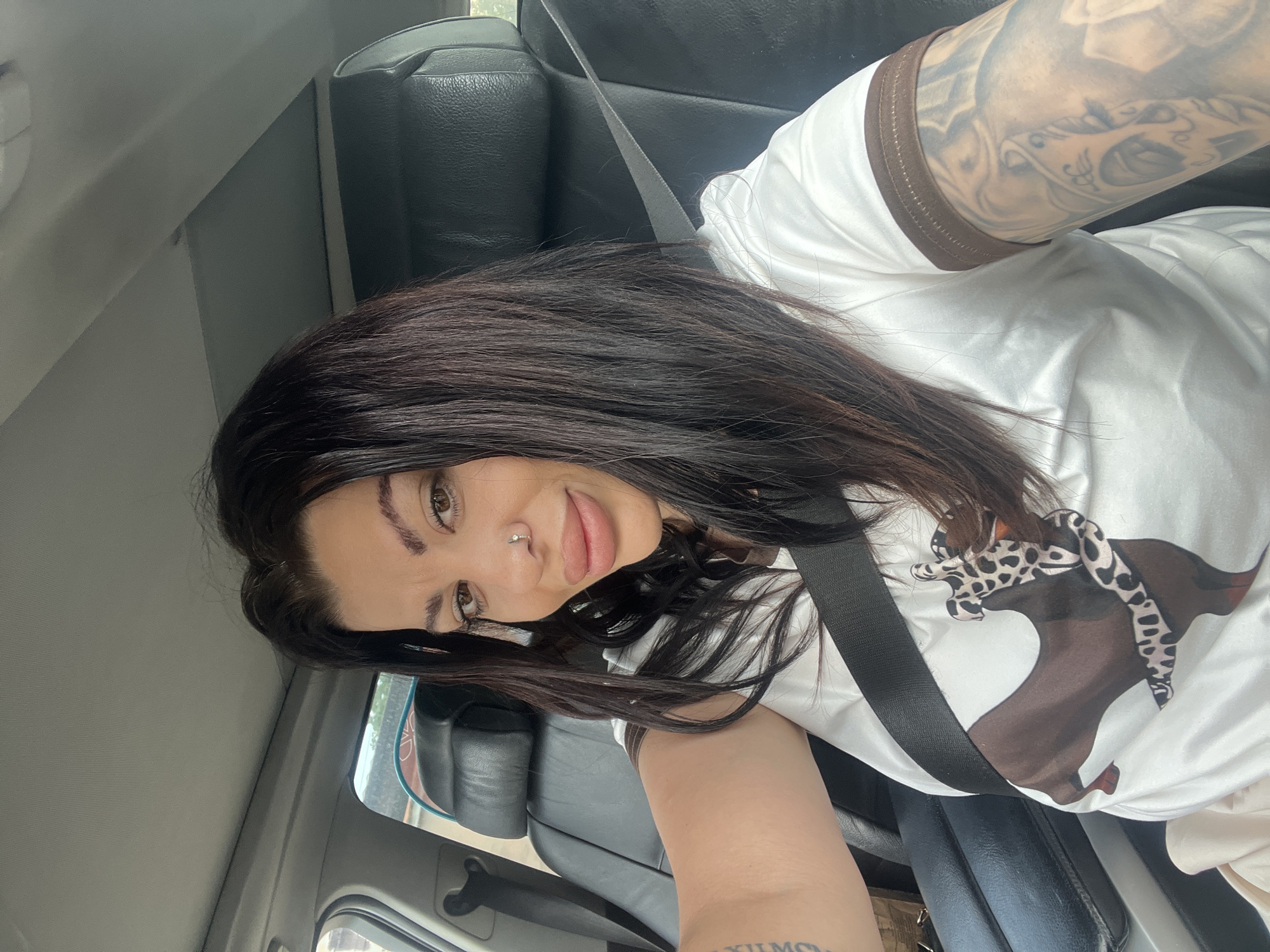 A medium shot captures a woman with long, dark hair and noticeable makeup, including prominent lip filler, wearing a white t-shirt with a graphic print of a bull and a snake, and a seatbelt, as she sits in the driver's seat of a car. The interior of the car, featuring black leather seats and a sunroof, is visible. The image appears to be taken during daylight. The location is identified as Kalekovets, Bulgaria.