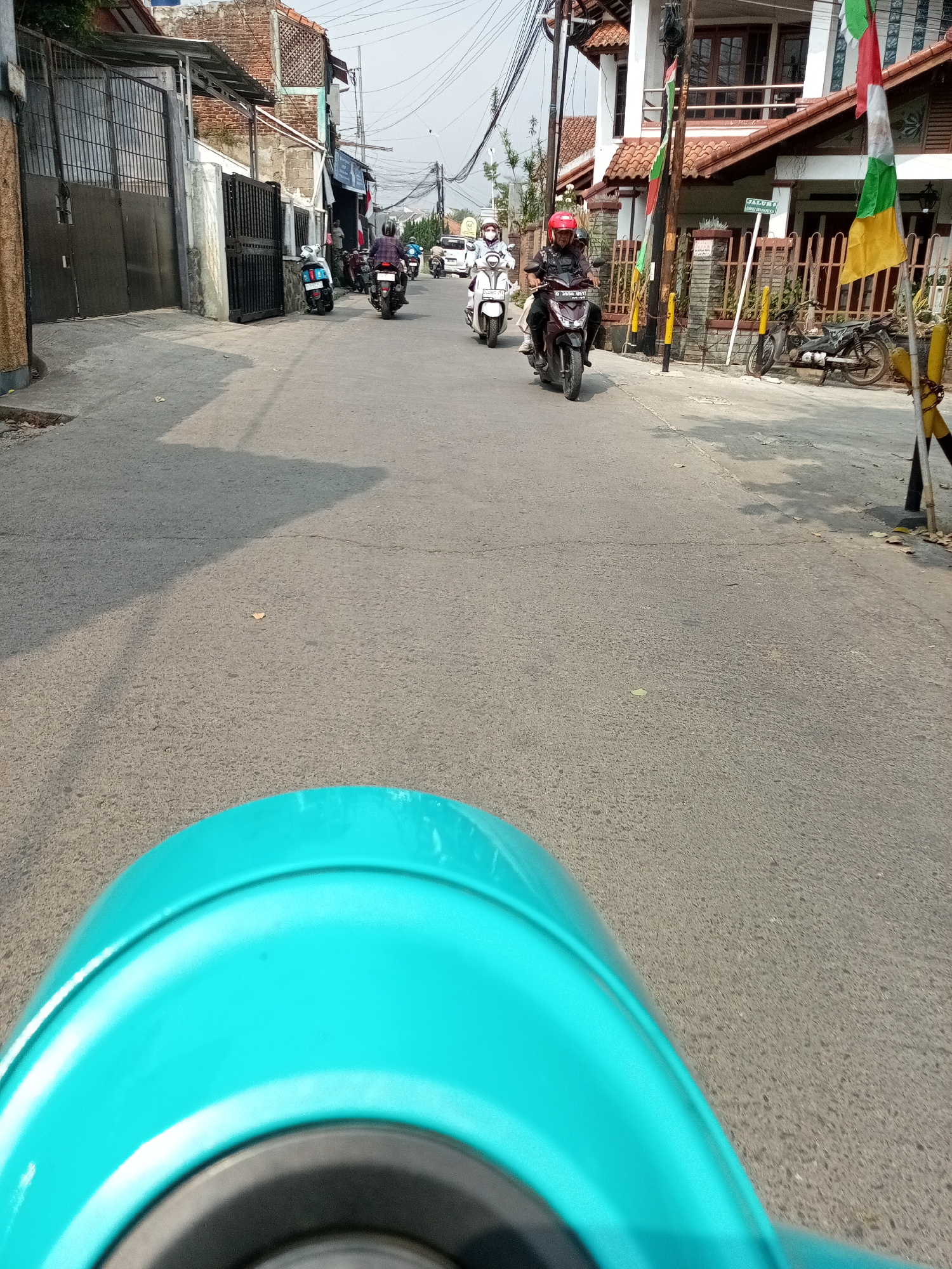 The image captures a street scene in Cihanjuang, Indonesia, viewed from the perspective of someone riding a teal scooter. The road is paved and appears to be a residential or commercial street, with buildings lining both sides. Several other motorcycles and scooters are present on the road. In the foreground, the teal body of the scooter being ridden is visible. The middle ground shows a procession of motorcycles moving along the street. Some riders are wearing helmets and face masks. The background features buildings with tiled roofs, some with visible flags. Utility poles with numerous wires are also prominent. The lighting suggests it is daytime, with shadows cast on the road. The environment is characteristic of a typical Indonesian urban or semi-urban setting.
