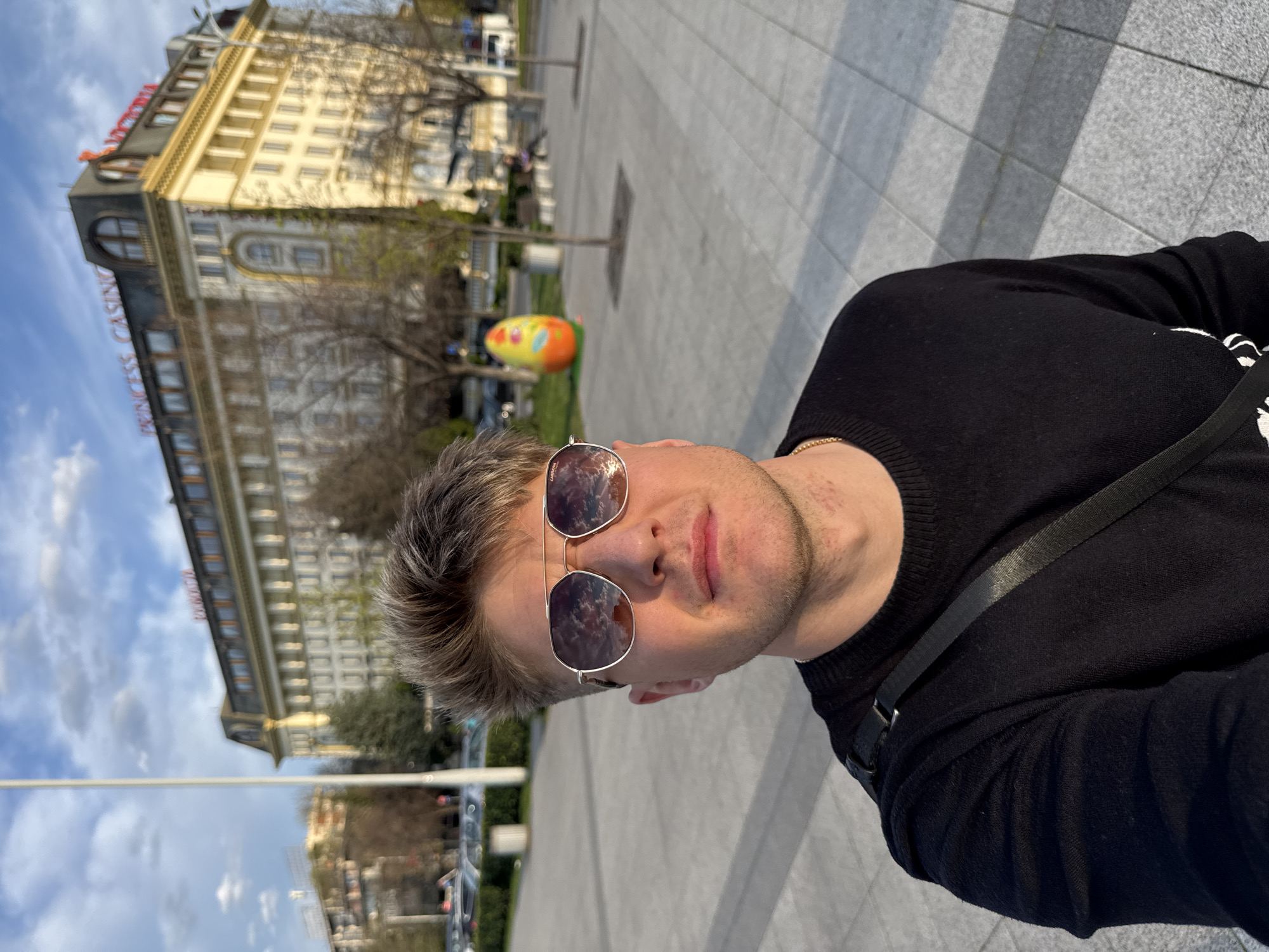 A medium shot of a man in sunglasses, wearing a black sweater and a black shoulder bag, is set against a background of a city scene, Plovdiv, Bulgaria. The man has blond hair. Reflected in his sunglasses are clouds. Behind him stands a large, ornate building with the words "PRINCESS CASINO" on its side. The building appears to be a hotel or casino with multiple stories. In the background, there's a decorated Easter egg. The ground is paved with stone tiles. The scene is bathed in natural light.