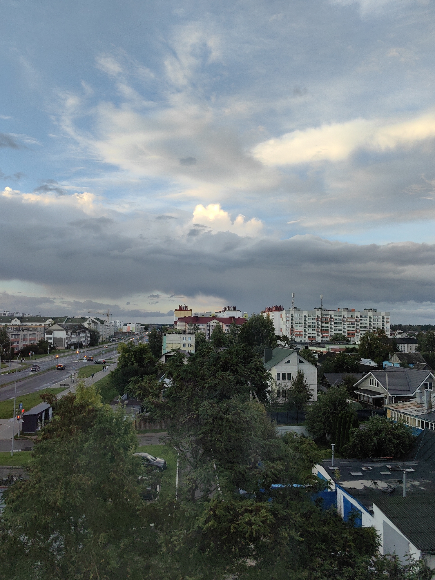 This aerial photograph captures a street scene in Fanipal, Belarus, with a focus on urban and suburban elements under a dramatic sky. A multi-lane road runs horizontally across the image, with several cars driving on it. Pedestrians can be seen on the sidewalk.
To the left of the road, a bus stop shelter is visible. Beyond the road, the cityscape unfolds. A long, multi-story apartment building with numerous windows and balconies dominates the middle ground on the right side. Smaller residential houses, some with pitched roofs, are interspersed with trees and vegetation throughout the scene.
The sky above is a prominent feature, with heavy, dark gray clouds occupying the upper half of the frame, suggesting impending or ongoing weather. Lighter clouds and patches of blue sky are visible towards the top, with some areas illuminated by sunlight. The overall lighting suggests it may be late afternoon or early evening. Green trees, some with dense foliage, are present in the foreground and scattered throughout the urban landscape, softening the visual impact of the buildings and infrastructure.