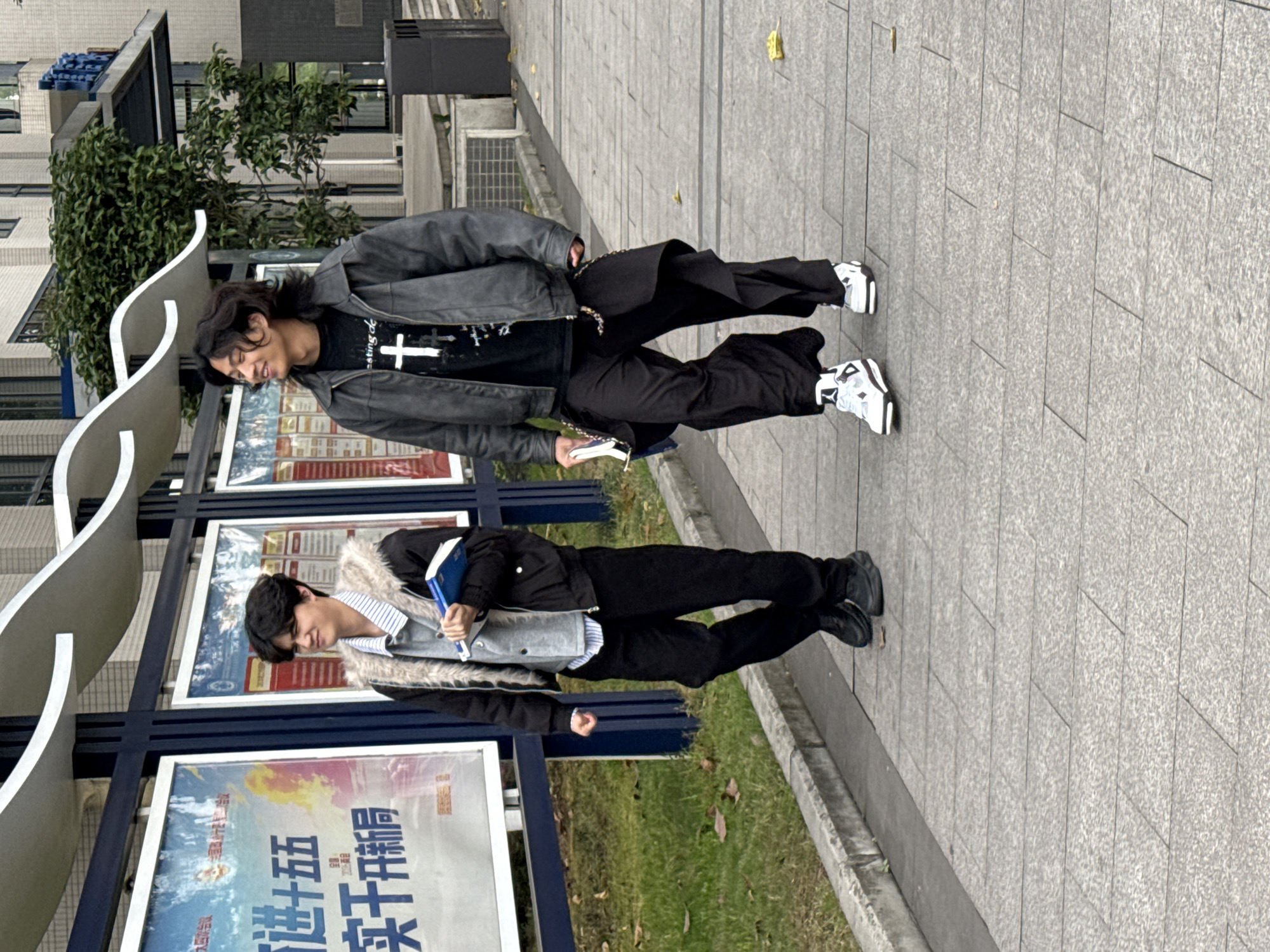 Two people are standing on a paved surface next to a bus stop structure. The bus stop has advertisements posted. The person on the left is wearing a black jacket with a fur collar, black pants, and black shoes, and is holding a blue book. The person on the right is wearing a gray jacket, a black T-shirt with a cross, black pants, and white sneakers, and is holding a smartphone. The scene is in Jiangjin, China.