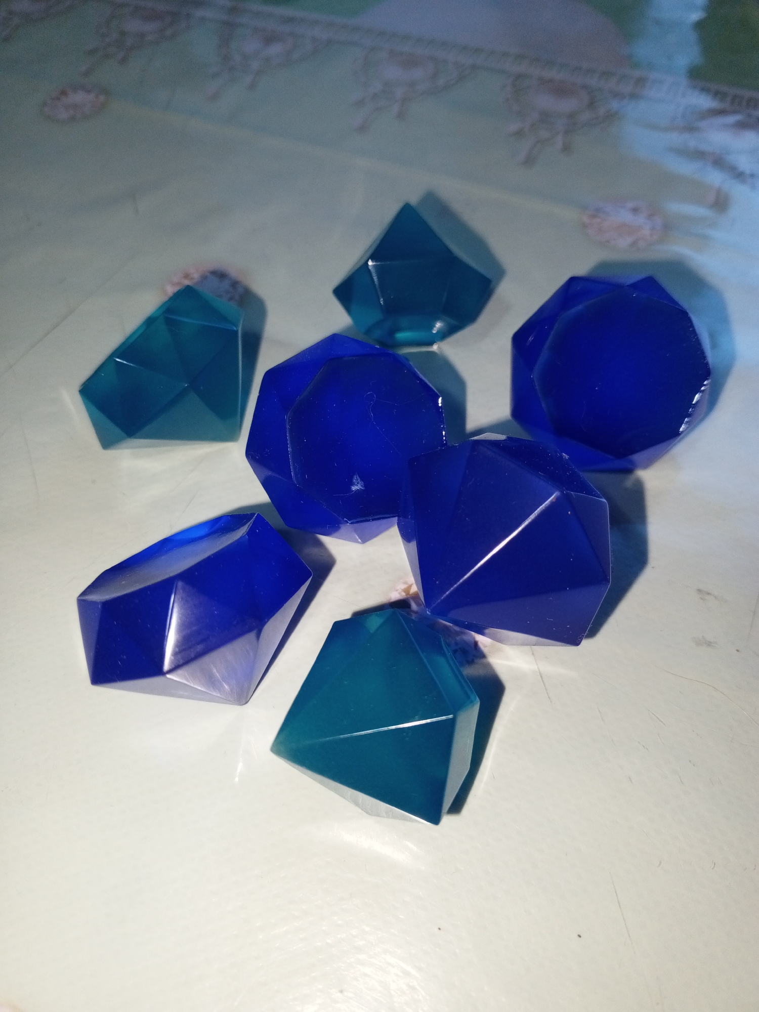 The image shows a collection of seven faceted, jewel-shaped objects scattered on a light-colored, textured surface. The objects appear to be made of translucent resin or glass, with some being a deep blue and others a turquoise or teal color. They are arranged in a loose cluster, with no discernible pattern. The lighting creates strong highlights and shadows on the surfaces of the objects, emphasizing their geometric shapes. In the background, a tablecloth or patterned surface is visible, with a faint repeating design. The overall impression is of decorative items or perhaps game pieces. There is no text visible in the image. The scene appears to be indoors, with artificial lighting. The context provided indicates the location is Saint Petersburg, Russia, but there are no specific visual cues in the image to confirm this.