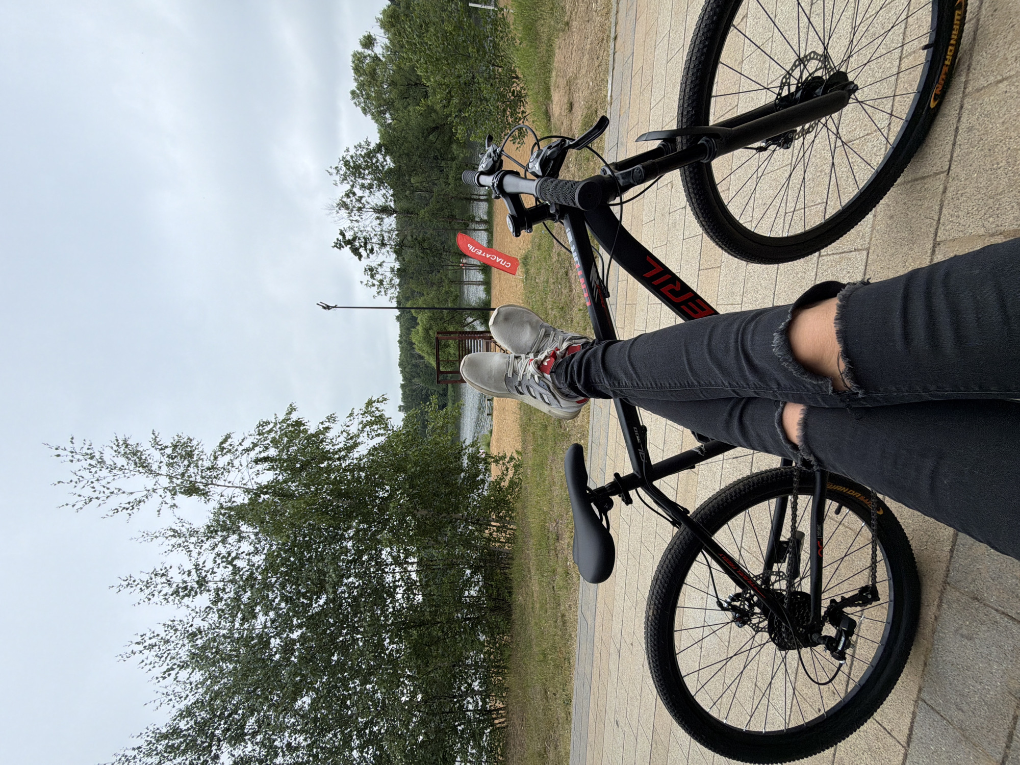 The image depicts a person's legs crossed in front of a black bicycle. The person is wearing black ripped jeans and gray sneakers. The bicycle has a red frame with black lettering and is parked on a paved walkway. In the background, there is a body of water, trees, and a red flag with Russian text that translates to "RESCUE". The overall scene suggests an outdoor recreational area, possibly a park or a waterfront in Malakhovka, Russia. The sky is overcast, indicating a cloudy day.