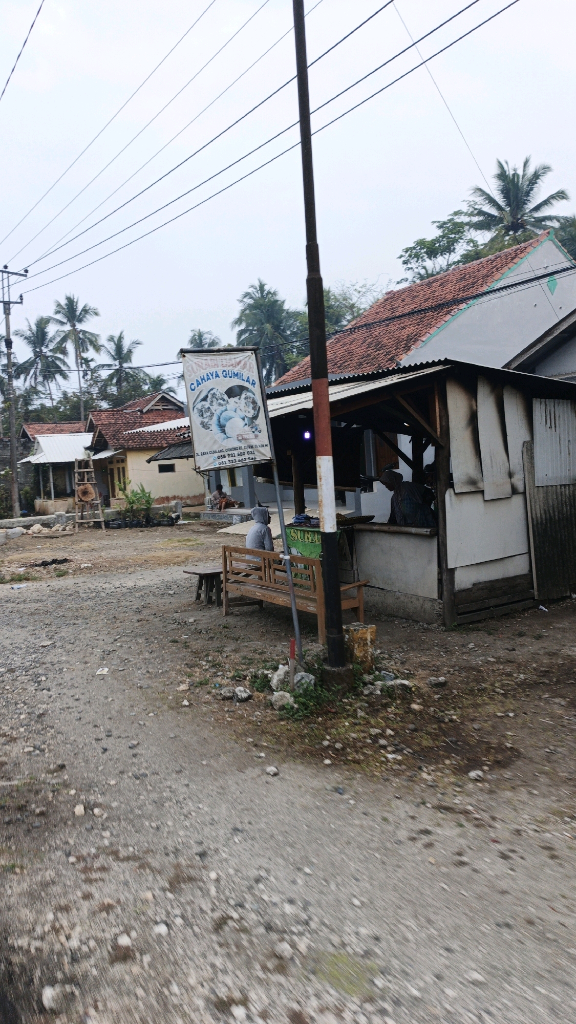 In Sukaresik, Indonesia, on a cloudy day, a roadside stall operates next to a dusty, gravelly path. A wooden bench sits unoccupied in front of the stall. Behind the bench, a man in a grey hooded jacket is seated. The stall itself has an open-front structure with a corrugated metal roof and a simple wooden counter. Inside, two individuals are visible, one in a blue shirt and the other in a grey shirt, seemingly engaged in conversation or work. Above them, a sign for "CAHAYA GUMILAR" displays images of eggs and text. The surrounding environment features several modest houses with orange tile roofs, palm trees, and utility poles with wires overhead, indicating a rural or semi-urban setting.