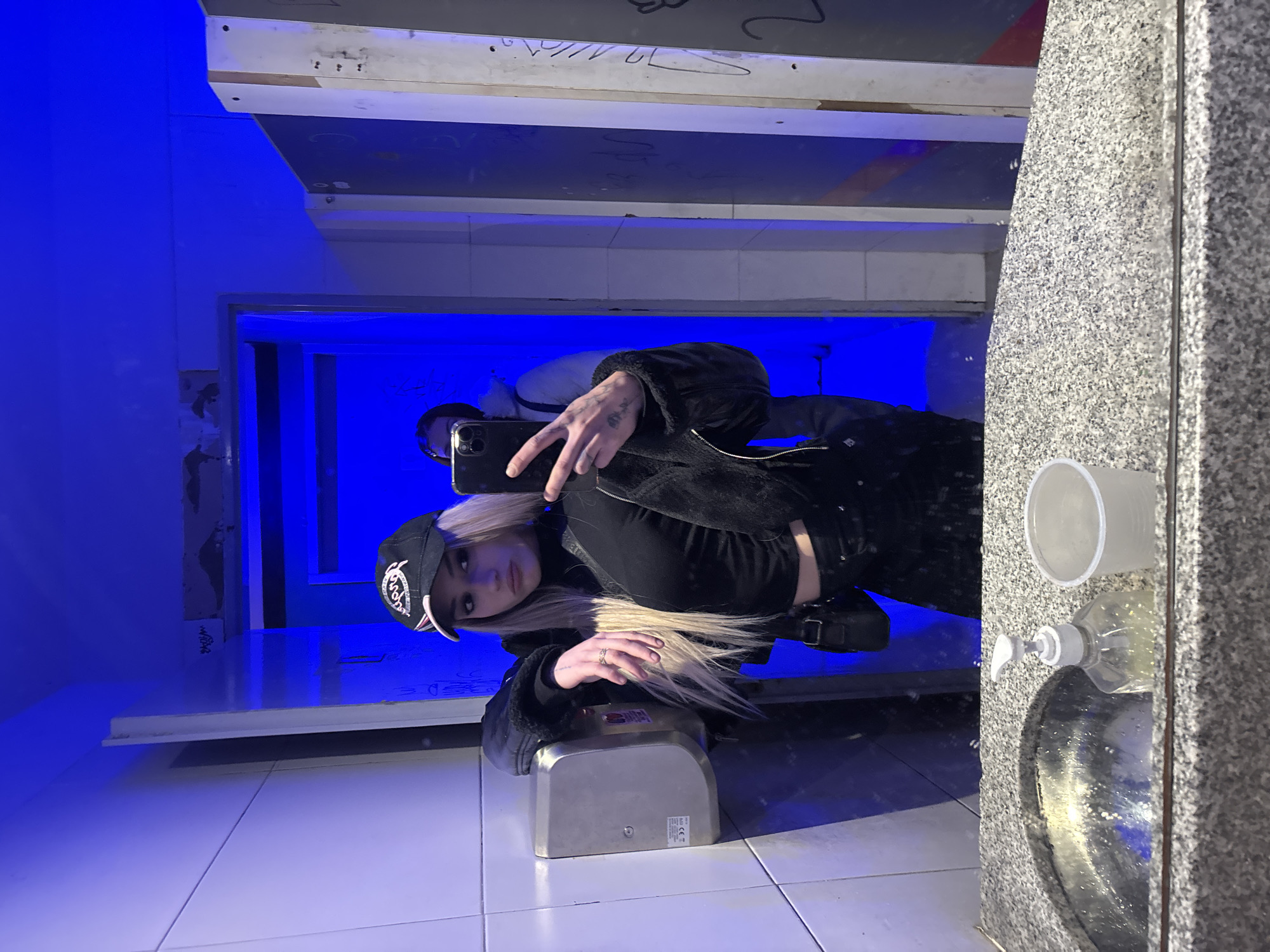 A young adult female is taking a selfie in what appears to be a public restroom. The restroom has blue lighting and graffiti on the walls. The female is wearing a black jacket, black top, black pants, and a baseball cap. She is holding a smartphone in her left hand. The restroom has a sink and soap dispenser. The photo was taken at an angle and has been rotated, but no evidence indicates that the photo was taken in Buenos Aires, Argentina.