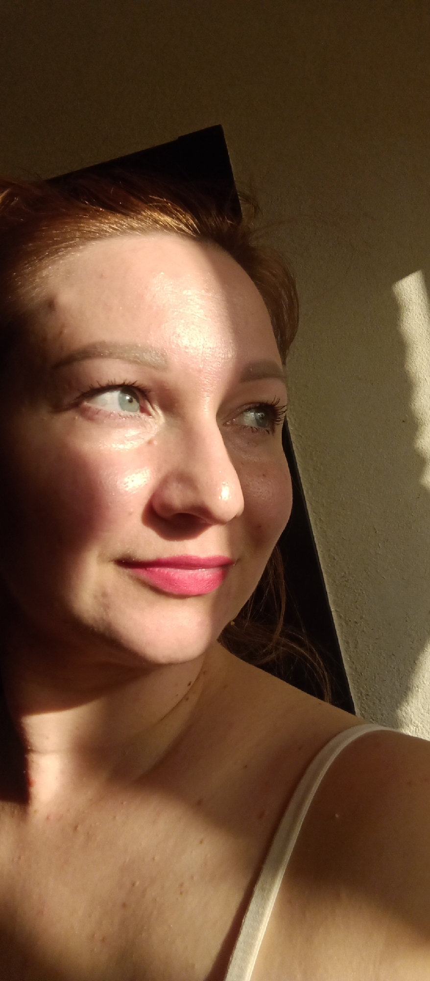 A red-haired woman is captured in a close-up, side-view portrait with a focus on her face. She has fair skin, with light freckling on her face and chest. Her lips are colored pink and she has light eye makeup. Light from an unseen source casts strong shadows across her face, leaving stark contrasts. Part of the background consists of a wall with the shadow of an object. A sliver of what appears to be a dark rectangular object is also visible to the left of her face. Her shoulder and a small portion of her white tank top are visible. The location and city are unknown.