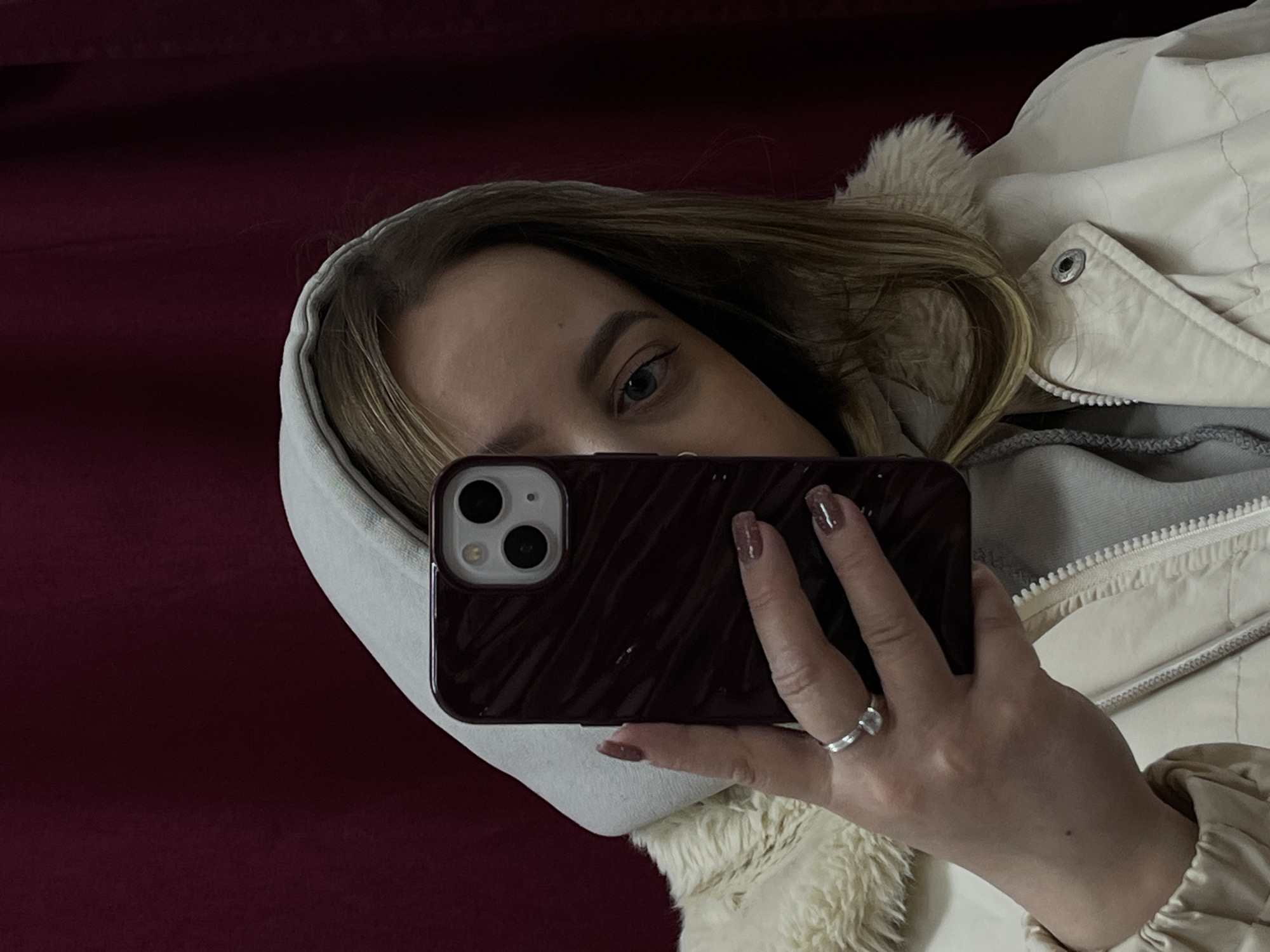 A woman is taking a selfie with a smartphone. She is wearing a hooded jacket. The background is a maroon color. There is no evidence to suggest that the image was taken in Novopokrovskaya, Russia.