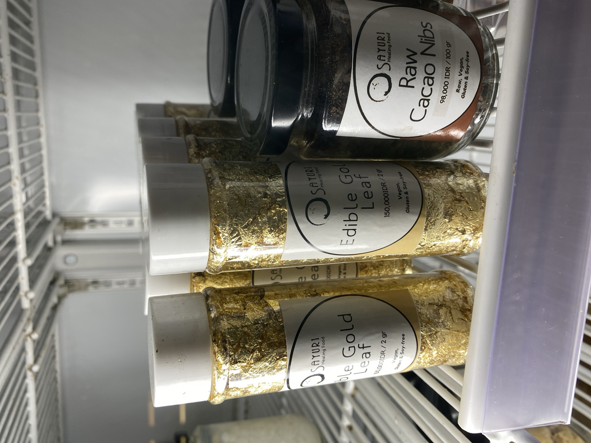 The image shows several jars and tubes of food products on a shelf, presumably in a shop or storage area. The foreground features tubes filled with what appears to be gold leaf, labeled "Edible Gold Leaf" with the brand name "SAYURI Healing Food". The tubes are white-capped and contain small pieces of gold leaf visible through the clear container. The label indicates a price of "150,000 IDR/2 gr" and notes that the product is vegan, gluten, and soy-free. Above these tubes are glass jars containing "Raw Cacao Nibs," also branded as "SAYURI Healing Food." These jars have black lids and a label indicating a price of "98,000 IDR/100 gr," and similarly specifies that they are raw, vegan, gluten, and soy-free.
The products are situated on a wire shelf, which suggests they are likely inside a refrigerator or storage unit. The shelves and interior of the unit are visible, with a white plastic divider partially obscuring the jars.
Based on the brand "SAYURI Healing Food", the location is the Ubud District, Indonesia.