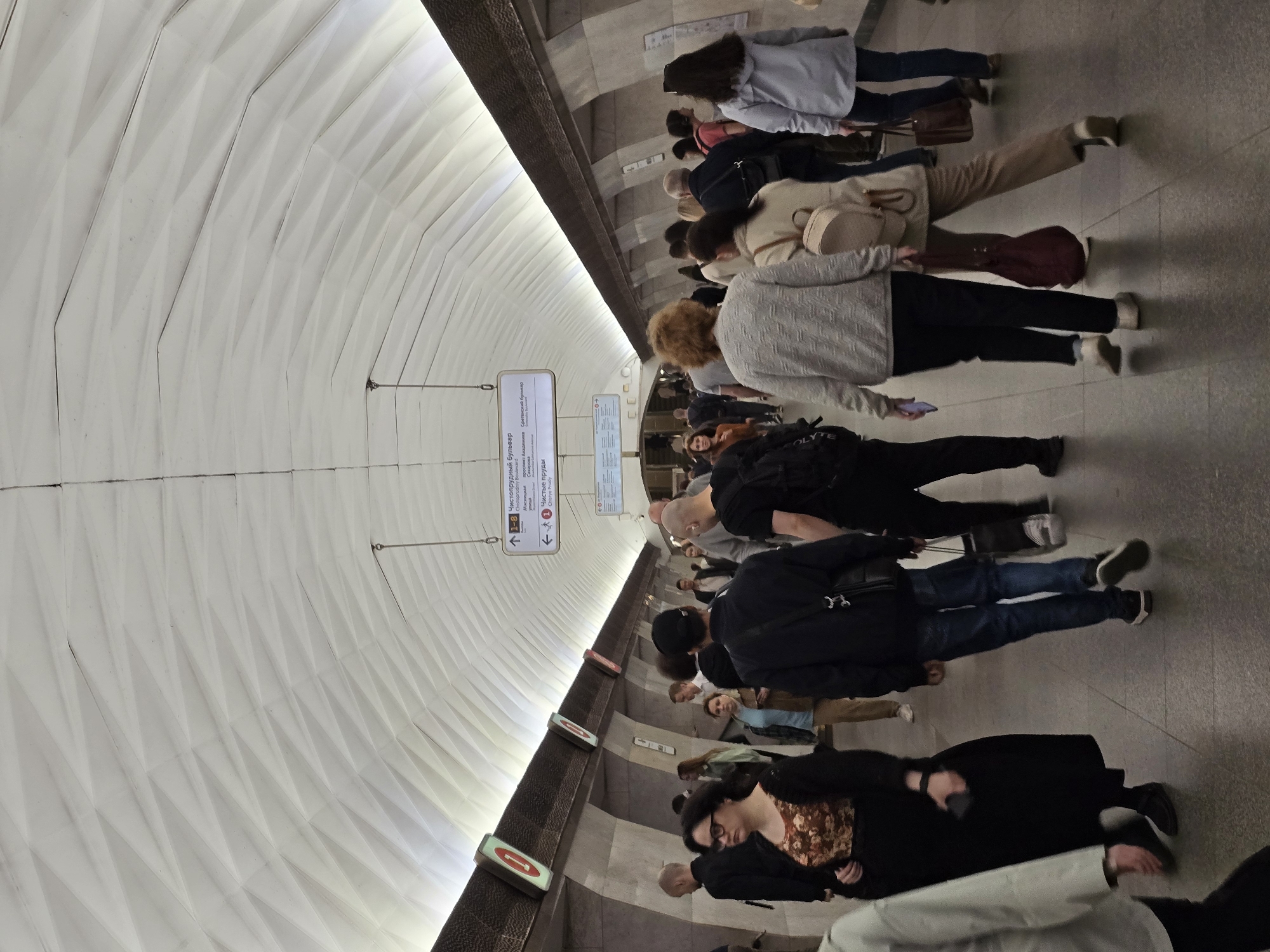 The image captures a scene within the Moscow Metro, Russia, specifically at the Chistye Prudy station. A group of pedestrians are observed walking through the station, their clothing varied, but predominantly dark colors. Directional signs are visible, indicating directions to "Chistoprudniy Boulevard", "Сретенский бульвар" (Sretensky Boulevard) and "Чистые пруды" (Chistye Prudy). The station's distinctive white geometric wall cladding is prominently displayed.