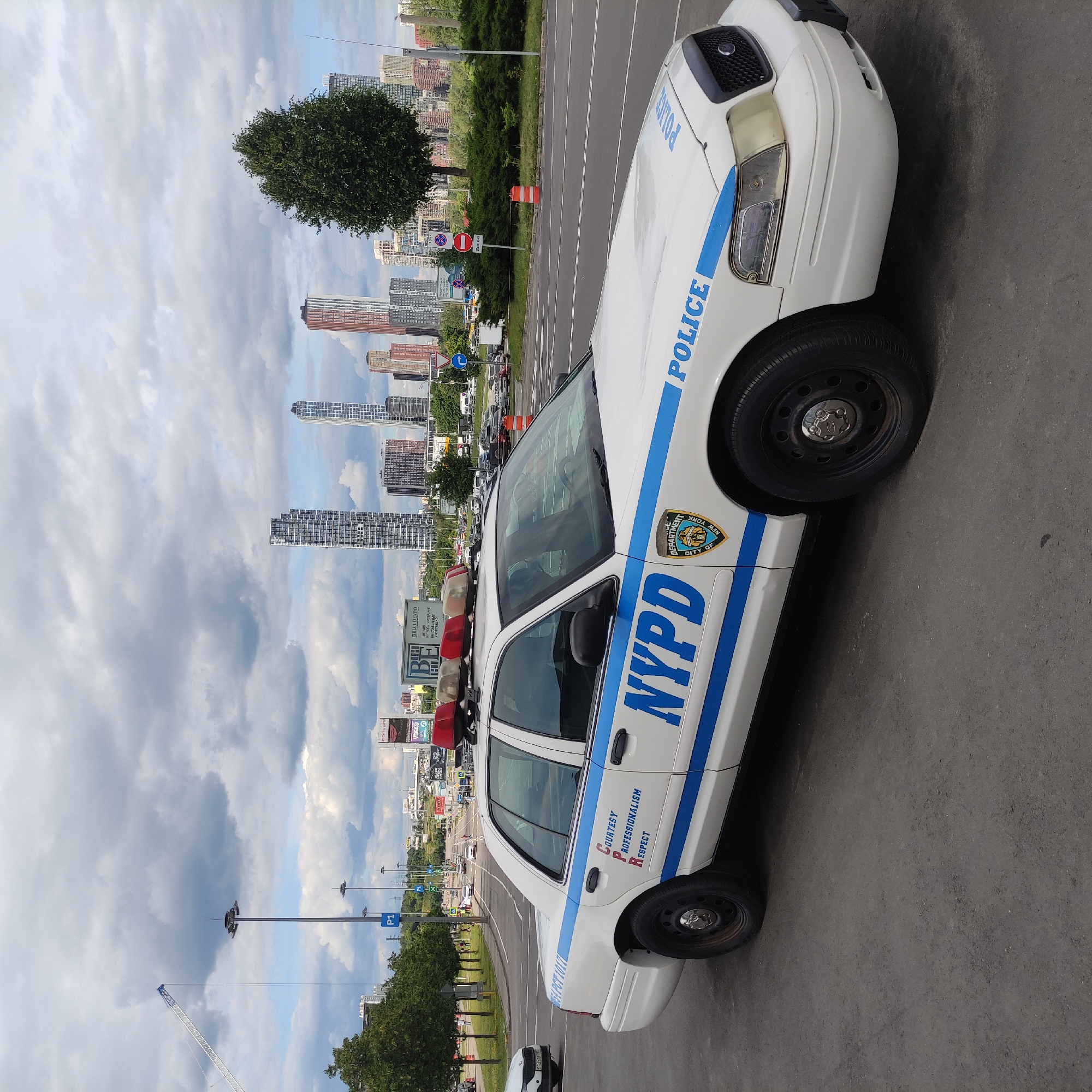 A New York City Police Department (NYPD) patrol car is parked on a street with a cityscape in the background. The car is white with blue stripes and the letters "NYPD" are visible on the side. A police shield is also present on the car. The cityscape features numerous tall buildings, some appearing to be under construction. Trees and a cloudy sky complete the scene. The image appears to be taken during the daytime. Signs are visible in the background, but the text is not legible. The location is identified as Khimki, Russia.