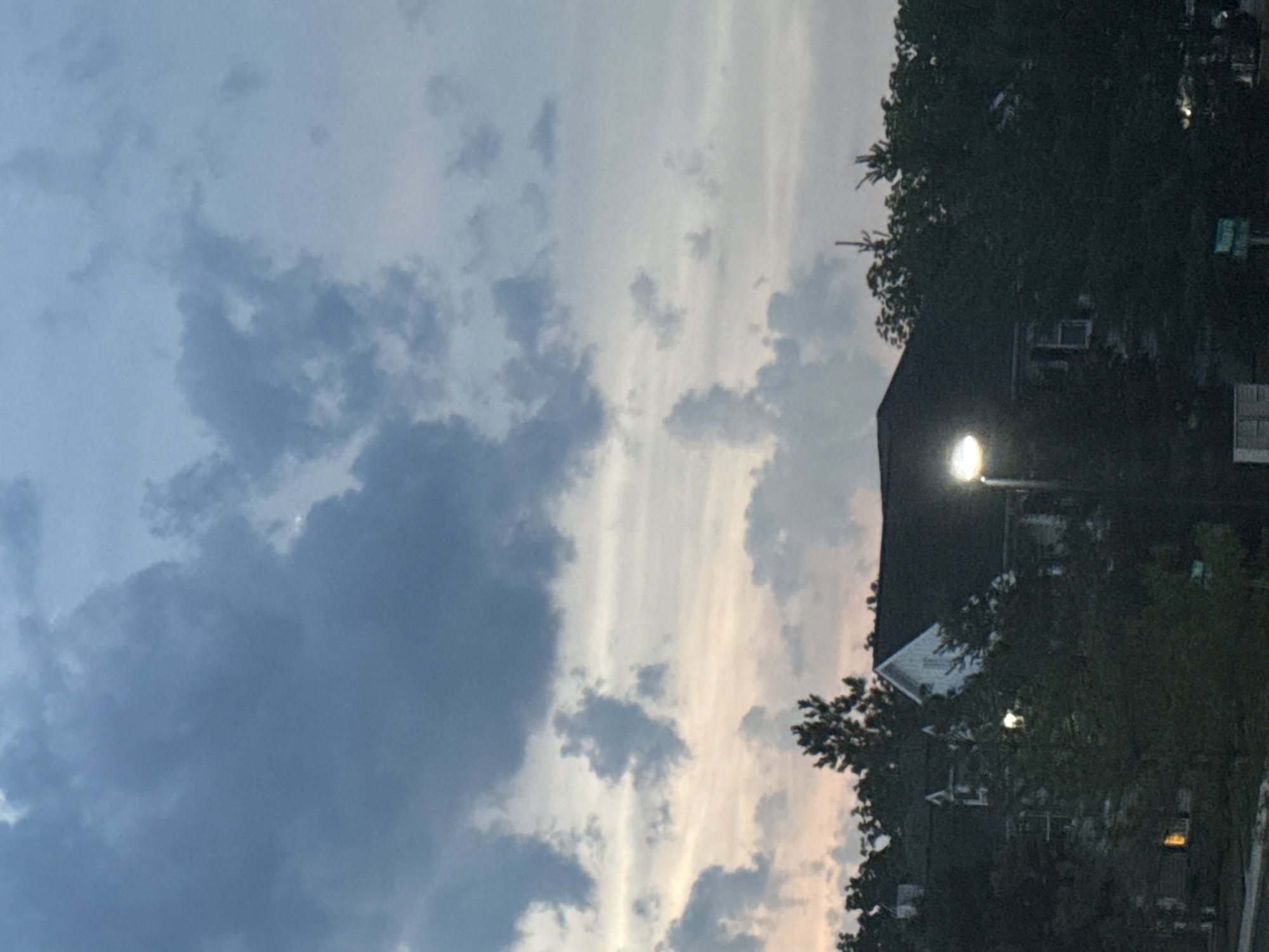 The image captures a dusky sky filled with dark, heavy clouds and lighter, wispy formations. The soft glow of sunset is visible near the horizon, tinging some of the clouds with pale pink and orange. Below the sky, a dark silhouette of trees and vegetation dominates the lower half of the frame. Partially obscured by the foliage are the dark roofs and white gables of houses. A streetlamp with a bright, glowing light is prominently positioned on the right side, illuminating the immediate surroundings. The scene appears to be a residential area, likely in a suburban or rural setting, as indicated by the houses and abundant trees. The overall atmosphere is serene and quiet, characteristic of twilight. The location is identified as Allen Township, United States of America.