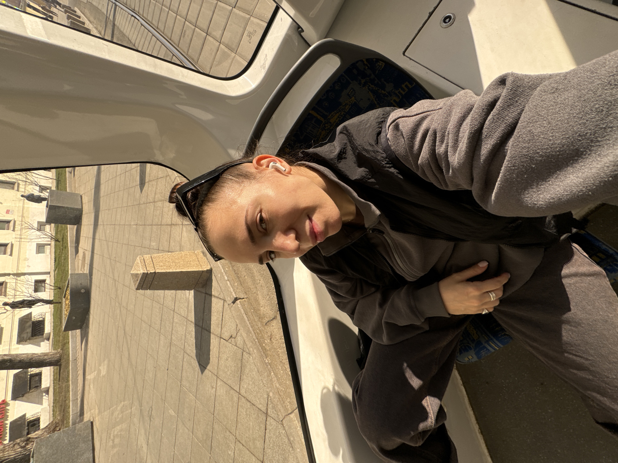 A woman is in a train in Moscow, Russia. She's wearing a grey sweatshirt and a black vest. Outside the window are buildings, trees, and paved walkways. There are also tram tracks visible outside the window. The woman is wearing sunglasses on her head and has an earbud in her ear.