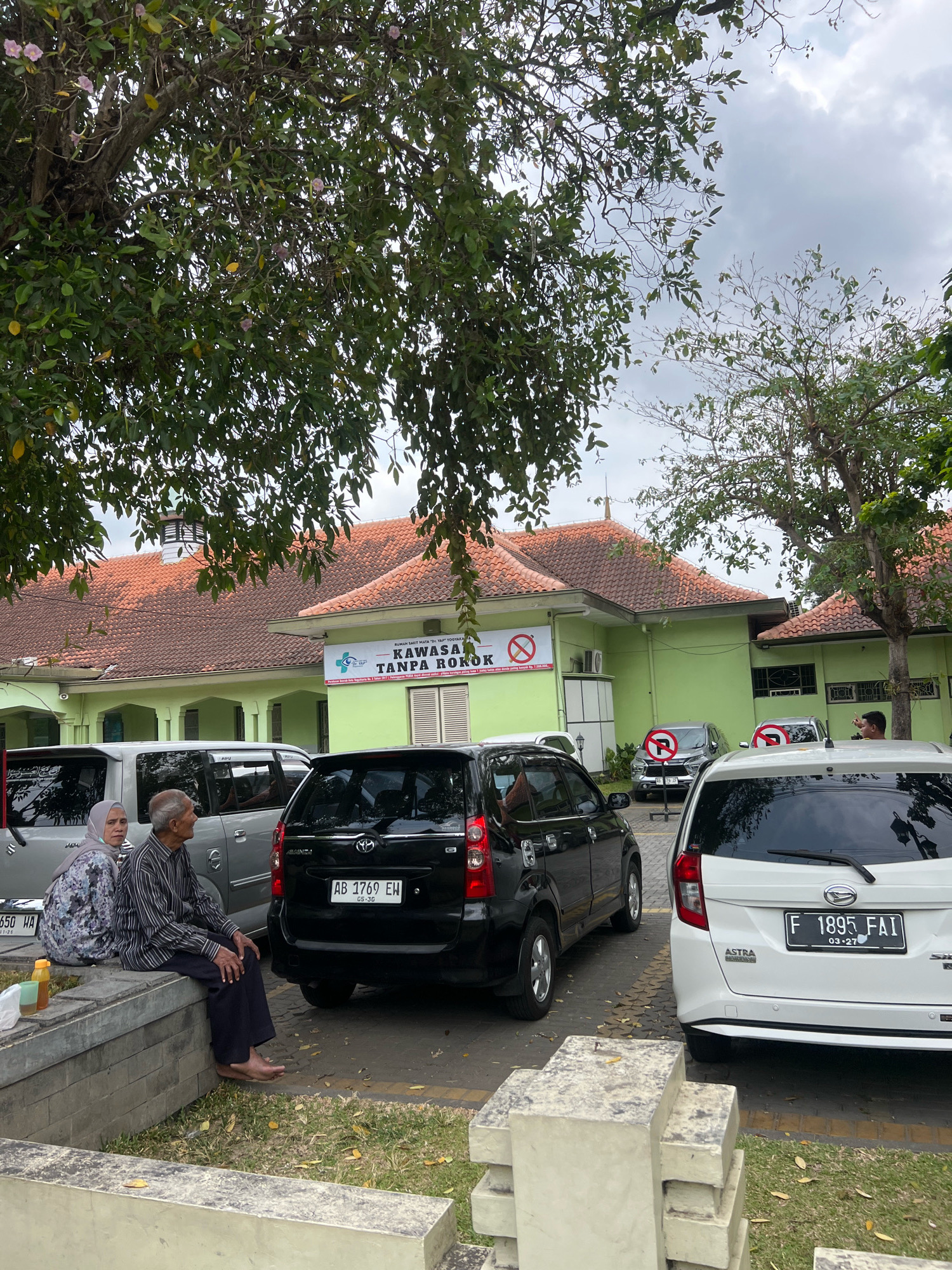 In front of a light green building with a terracotta tiled roof, several cars are parked in a lot. The building has a sign that reads "RUMAH SAKIT MATA "DR. YAP" YOGYAKARTA" and "KAWASAN TANPA ROKOK" which translates to "SMOKING FREE AREA". A couple, an elderly man and woman, are sitting on a concrete ledge in the foreground. The man is wearing a striped shirt and dark pants, and the woman is wearing a hijab and a patterned dress. They are facing away from the camera, looking towards the right of the image. A black minivan with a license plate reading "AB 1769 EW" is parked behind them. To the right of the minivan is a white car with the license plate "F 1895 FAI". Several trees with green leaves and some pink flowers are visible in the background. The sky is overcast.