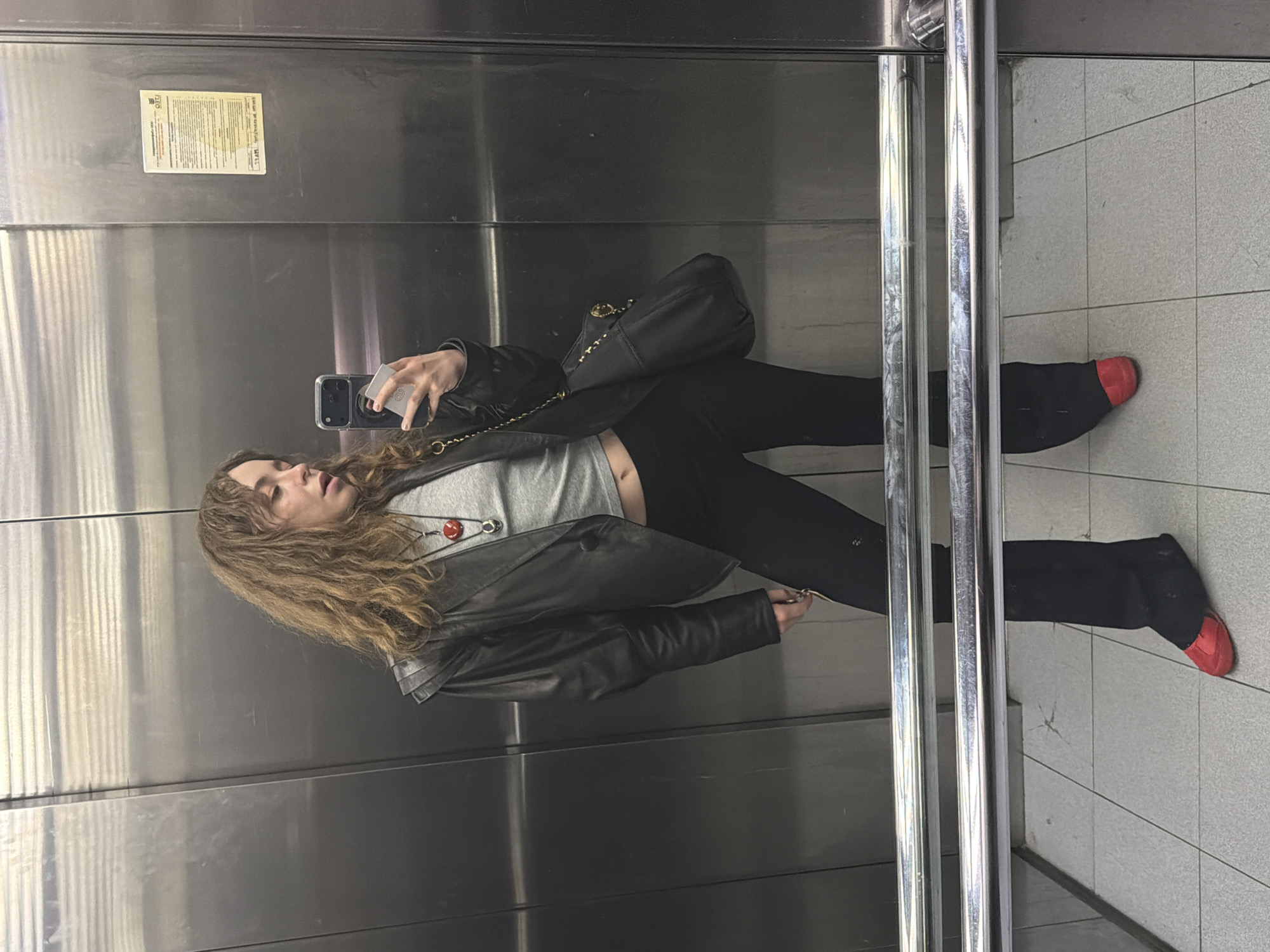 A woman stands in an elevator, taking a mirror selfie. She has curly brown hair and is wearing a grey t-shirt, black leather jacket, black leggings, and black socks with red toes. A black purse hangs over her shoulder. She is holding a phone in her left hand. The elevator has stainless steel walls and a notice posted on the top left. Through the elevator doors, a tiled floor is visible. The scene appears to be indoors. Based on context clues, the location of the image is likely to be Moscow, Russia.