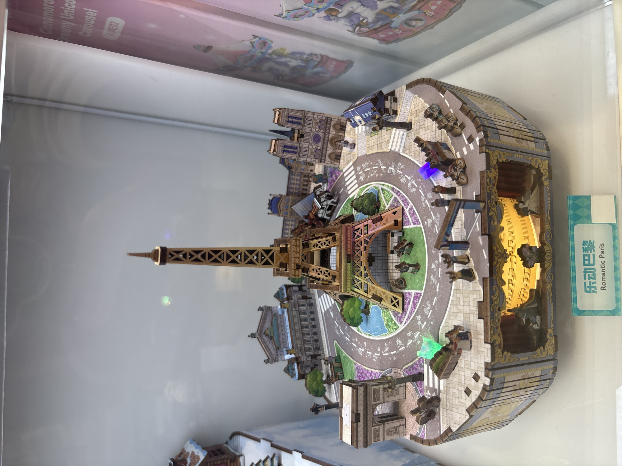 The image displays a detailed model of Paris, featuring iconic landmarks like the Eiffel Tower, Arc de Triomphe, and Notre Dame Cathedral. The model is intricately designed with miniature figures, trees, roads, and a small pond. The base of the model is decorated with ornate patterns and has a cutout that reveals a lit scene with figures and musical notes. A sign at the bottom reads "乐动巴黎 Romantic Paris". The model appears to be on display, possibly in a shop or museum, within a glass case. The background is a light gray, suggesting an indoor environment. There is no indication of the specific location in Nha Trang Ward, Vietnam, within the image itself.