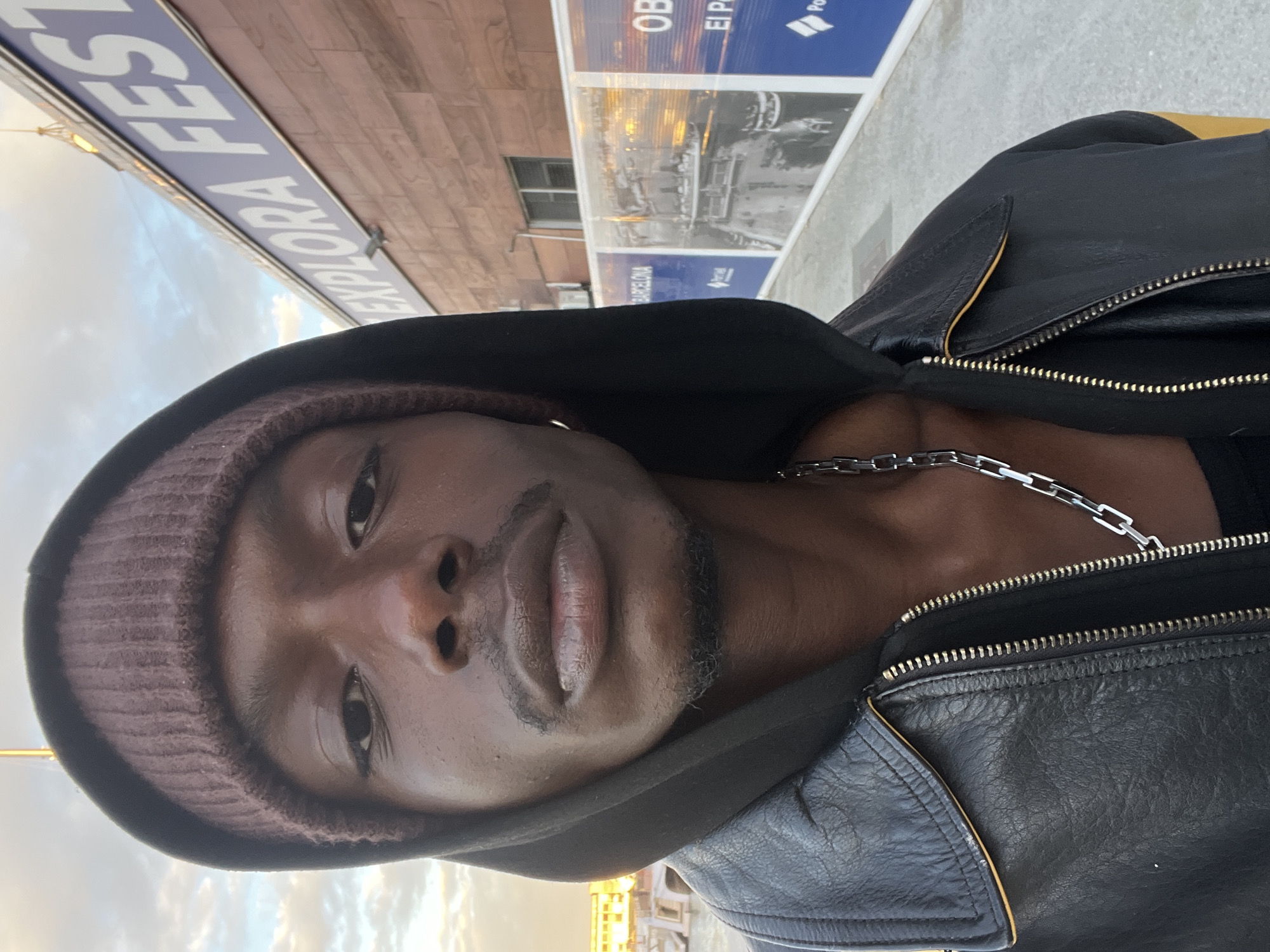 A medium shot captures a Black man in a brown beanie and hooded black leather jacket. He wears a silver chain necklace. Behind him is the Port Olímpic in Barcelona, Spain, with a billboard for an exhibit at El Born CCM titled "EXPLORA FEST."