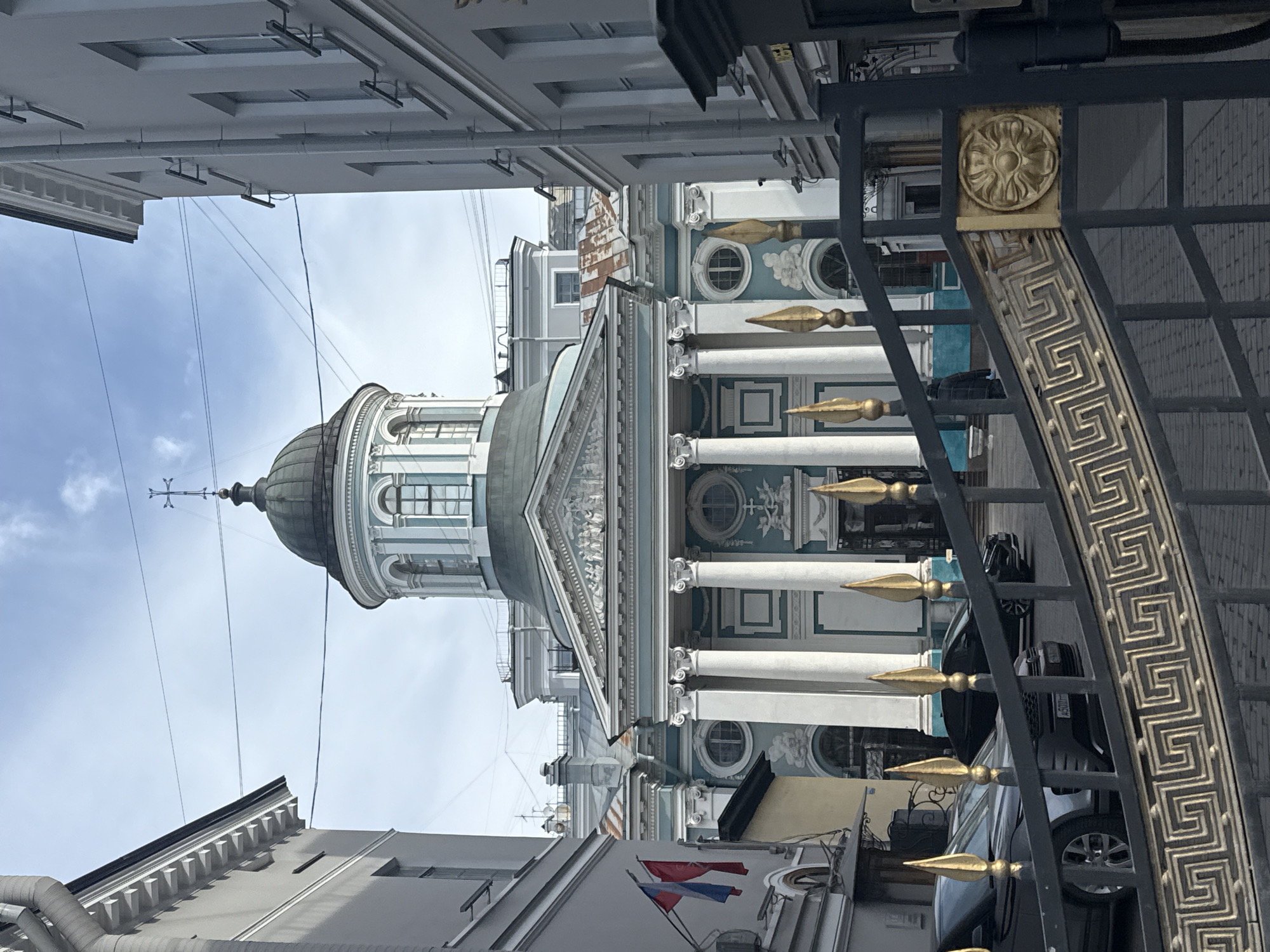 This image, when correctly oriented, presents a street-level view of a grand architectural structure in Saint Petersburg, Russia, seen partly through a decorative metal fence.
The central subject is a majestic building featuring a large, dark green dome topped with a spired cupola and a cross. The facade below the dome is light blue-green with prominent white architectural elements, including classical columns with ornate capitals, supporting a detailed pediment that appears to contain sculptural relief work. The building also features circular and rectangular windows with elaborate frames, and intricate decorative motifs along its structure.
In the foreground, a black metal fence or gate with striking gold accents partially obscures the view. This fence is adorned with a Greek key (meander) pattern in gold and a circular gold medallion featuring a floral design. Several gold, spear-like finials extend upwards from the fence, and some are also visible behind it.
Behind the fence, parts of at least two vehicles are visible, including a dark-colored SUV or crossover with a visible license plate segment reading "A 500".
To the left and right of the main building, other less ornate, light-colored buildings with windows and rooflines are visible, typical of an urban street. In the bottom left corner, on a smaller building, two small flags are displayed; one appears to be the Russian flag (red, white, and blue), while the other, also red, white, and blue, is less distinct.
The sky overhead is light blue with scattered white clouds, indicating clear or partly cloudy weather. Utility or electrical wires stretch across the upper part of the frame. The scene is illuminated by bright daylight, suggesting either morning or afternoon. No people or specific activities are depicted, focusing instead on the impressive architecture and urban environment.