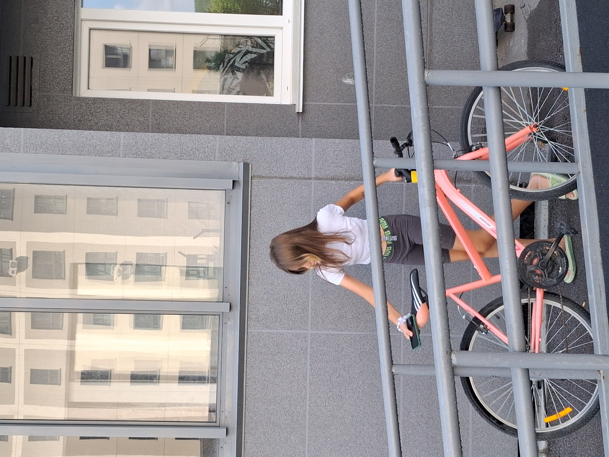 A young girl with long brown hair is standing next to a pink bicycle on a ramp in front of a modern building. She is wearing a white crop top and dark shorts. Her left hand is resting on the seat of the bicycle, and she is holding a smartphone in her right hand. The building has large windows that reflect the sky and other buildings. The ramp is made of metal and is part of the building's entrance. A skateboard is visible on the ground to the right of the bicycle. The image appears to have been taken during the day, under bright sunlight, suggesting a warm weather environment. The architecture and the general appearance of the building are consistent with contemporary urban design found in cities like Novosibirsk, Russia.