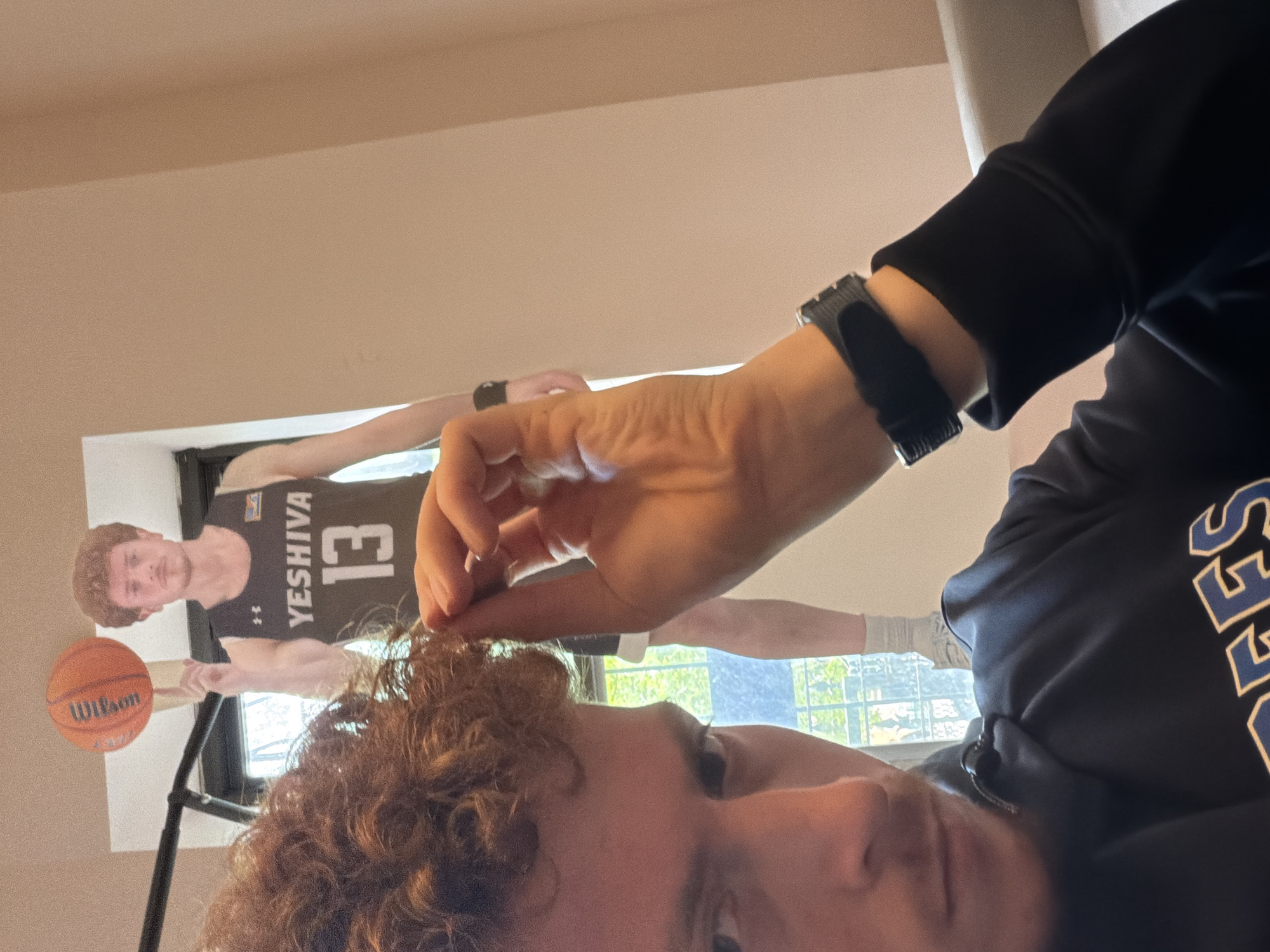 The image shows a person with curly reddish-brown hair holding a strand of their hair between their fingers. They are wearing a dark-colored sweatshirt with light-colored lettering. A black smartwatch is visible on their wrist. In the background, there is a poster of a basketball player wearing a Yeshiva jersey with the number 13. The player is holding a Wilson basketball on his finger, and there is an indoor basketball hoop behind him. Behind the player, a window shows a view of trees and sky. Assuming Yeshiva refers to Yeshiva University, the location is likely somewhere in New York City, United States of America.