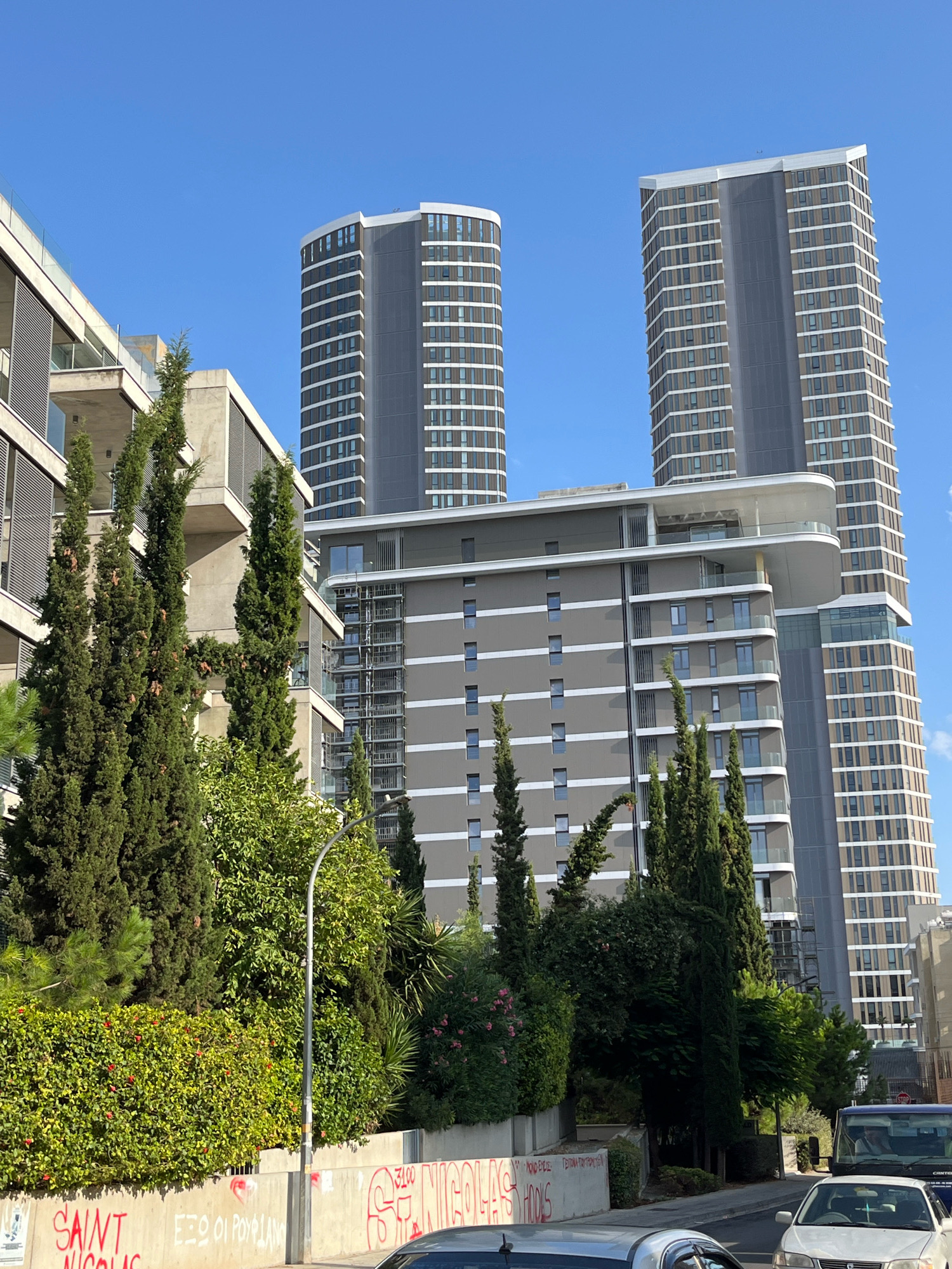The image captures a street scene in Limassol, Cyprus, with a clear blue sky overhead. In the background, two modern, tall apartment buildings dominate the skyline. The building on the right is a rectangular skyscraper with a facade of regularly spaced windows and vertical grey panels. The building to its left is also a high-rise, but with a curved facade and a similar window pattern. In front of these, a mid-rise building with a grey exterior features horizontal bands of white and numerous windows.
The foreground is occupied by a dense arrangement of evergreen trees, including tall cypress trees and a lush green hedge. Some pink flowers are visible amongst the foliage. A concrete wall runs along the bottom of the image, marked with red graffiti, including the words "SAINT NICOLAS". A street lamp with a curved pole stands near the wall. In the lower right corner, the front of a white car is visible, and behind it, the front of a dark blue van. The scene depicts an urban environment with a mix of contemporary architecture and natural elements.
