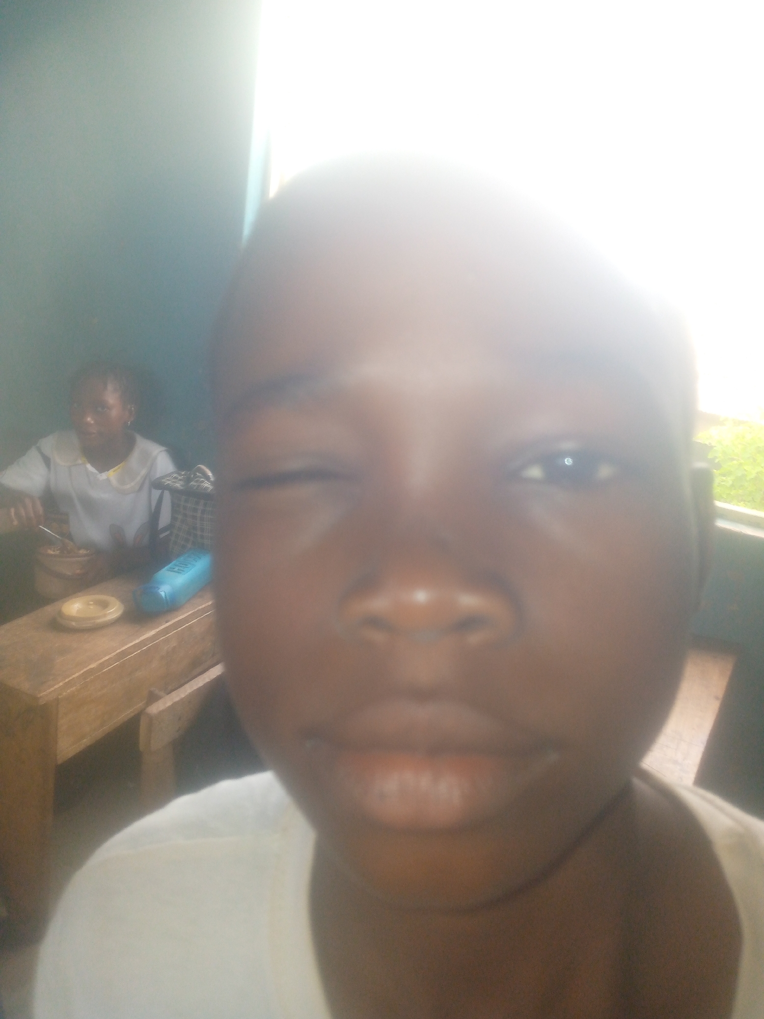 The image shows a close-up of a person with dark skin, winking at the camera. Behind them, slightly out of focus, another individual is sitting at a wooden table, possibly eating. A blue object and a dish are also visible on the table. The location is speculated to be Ikorodu, Nigeria, however, there are no specific identifiable characteristics of the city in the image itself. The lighting is bright, possibly from a window on the right. The overall scene appears casual, possibly indoors.