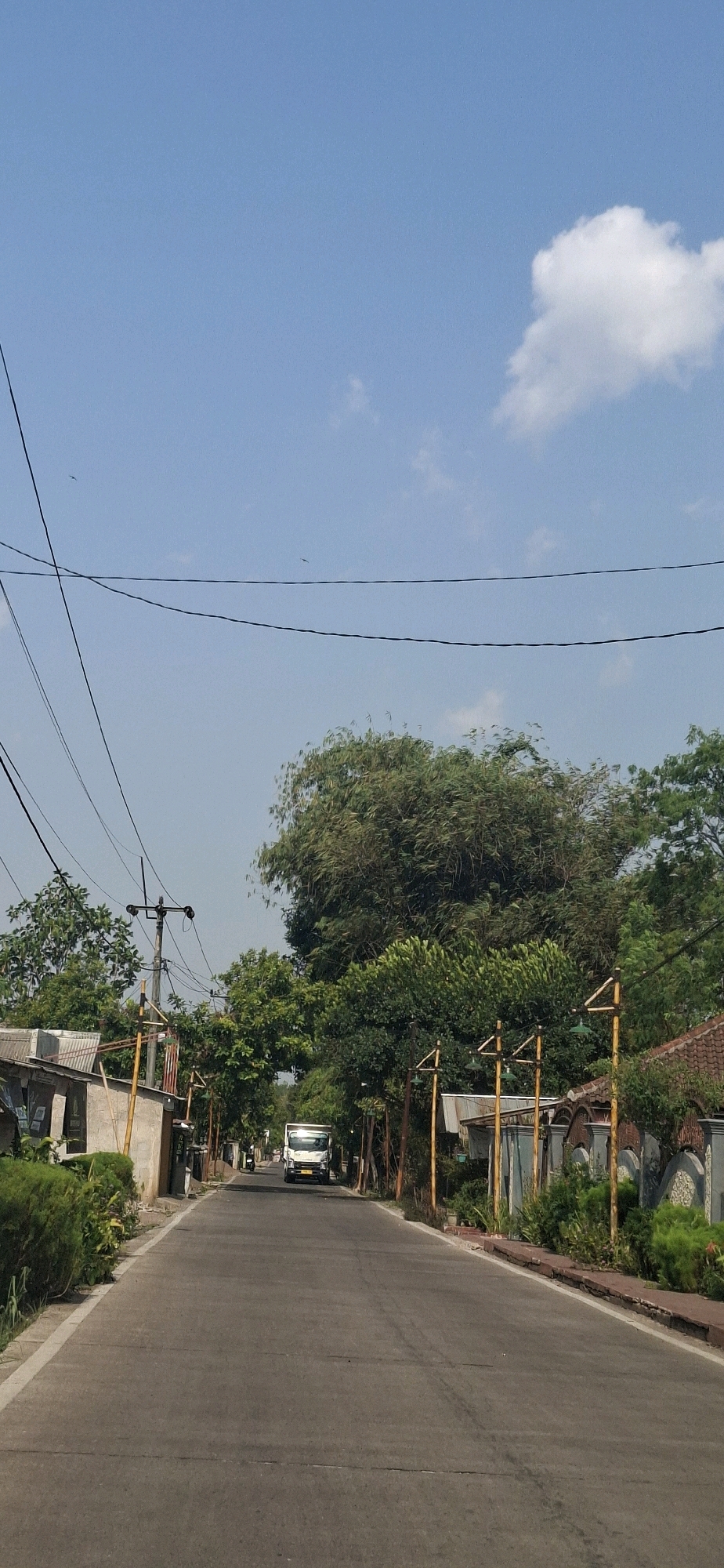 The image depicts a straight, paved road in Cibinong Hilir, Indonesia, lined with vegetation and residential structures. A white truck is visible in the distance, traveling down the road. Overhead, power lines crisscross the clear blue sky, with a few wispy clouds present. The presence of houses and the truck suggests a populated area, likely a residential or semi-rural neighborhood. The overall impression is of a typical day in a Indonesian town.