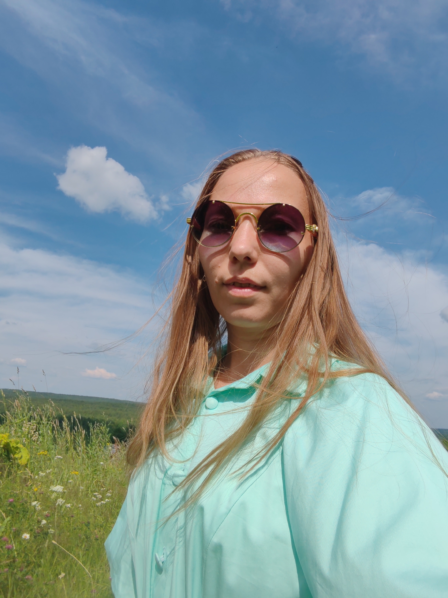 A young woman with long, blonde hair, wearing round, gold-rimmed sunglasses and a light turquoise shirt, stands outdoors. She is in the foreground, facing the camera. Behind her, a field of tall green grass and wildflowers stretches into the distance, leading to a rolling landscape of green hills under a bright blue sky with scattered white clouds. The scene appears to be in a rural or natural setting.