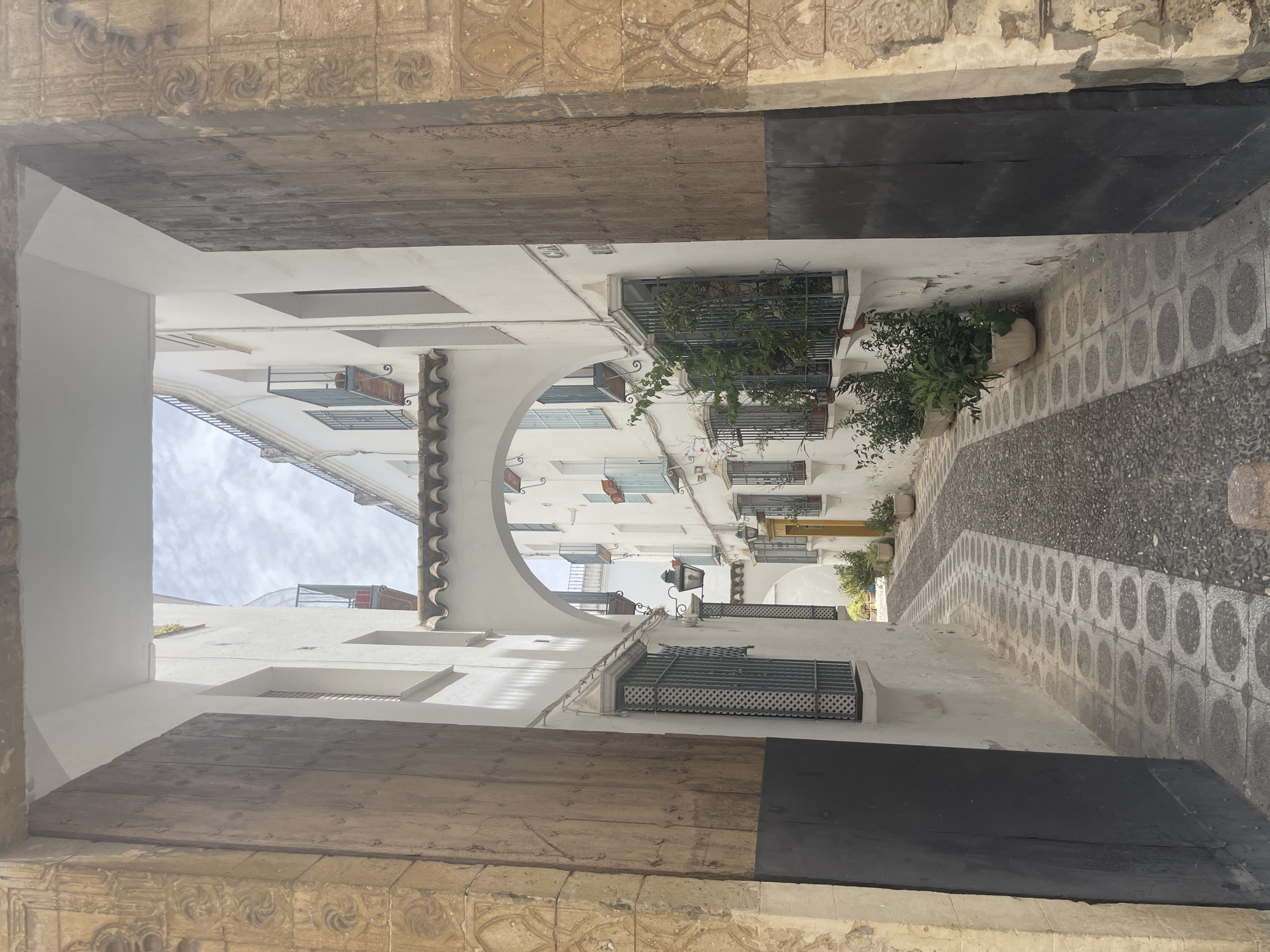 The image depicts a narrow, cobblestone alleyway in Córdoba, Spain, framed by large, ornate wooden doors and stone archways. The alley opens up to reveal traditional white-washed buildings with characteristic balconies adorned with plants and flowers. The architecture suggests a historic European city, likely with Moorish influences, common in Andalusia. The sky is cloudy, indicating moderate daylight. There are no people visible in the scene. The alley has a distinct patterned walkway, alternating between dark cobblestones and lighter circular inlays. The overall impression is one of charm and historical depth.