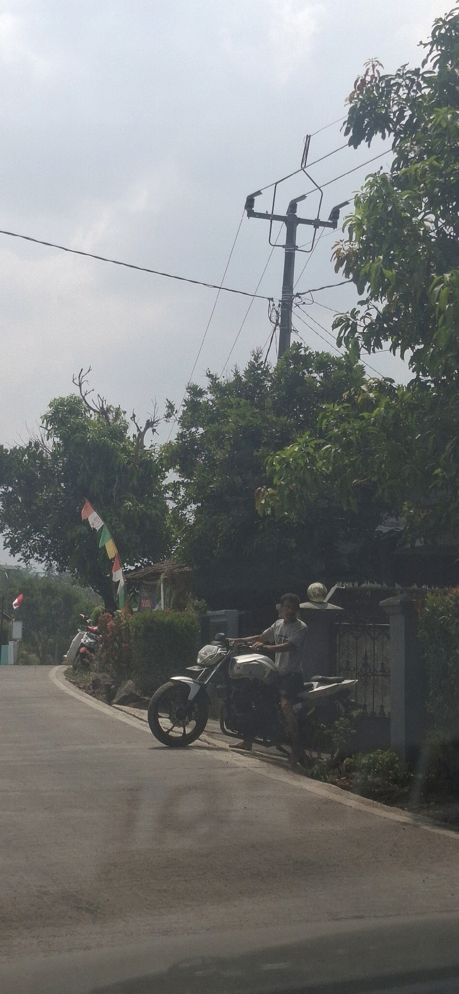 The image depicts a scene on a paved road in Cibinong Hilir, Indonesia. A young man is seated on a white and black motorcycle, parked on the side of the road. He is wearing a light grey t-shirt and dark shorts. The motorcycle is positioned to the right of the frame, facing towards the left. Behind the motorcycle, a weathered fence and lush green trees are visible. A flag, featuring red and white stripes along with green and yellow segments, is planted on the left side of the road, near a small structure. The sky is overcast with a muted grey hue. Power lines are visible spanning across the upper portion of the image, supported by a utility pole. The overall setting appears to be a rural or suburban area with vegetation and basic infrastructure.