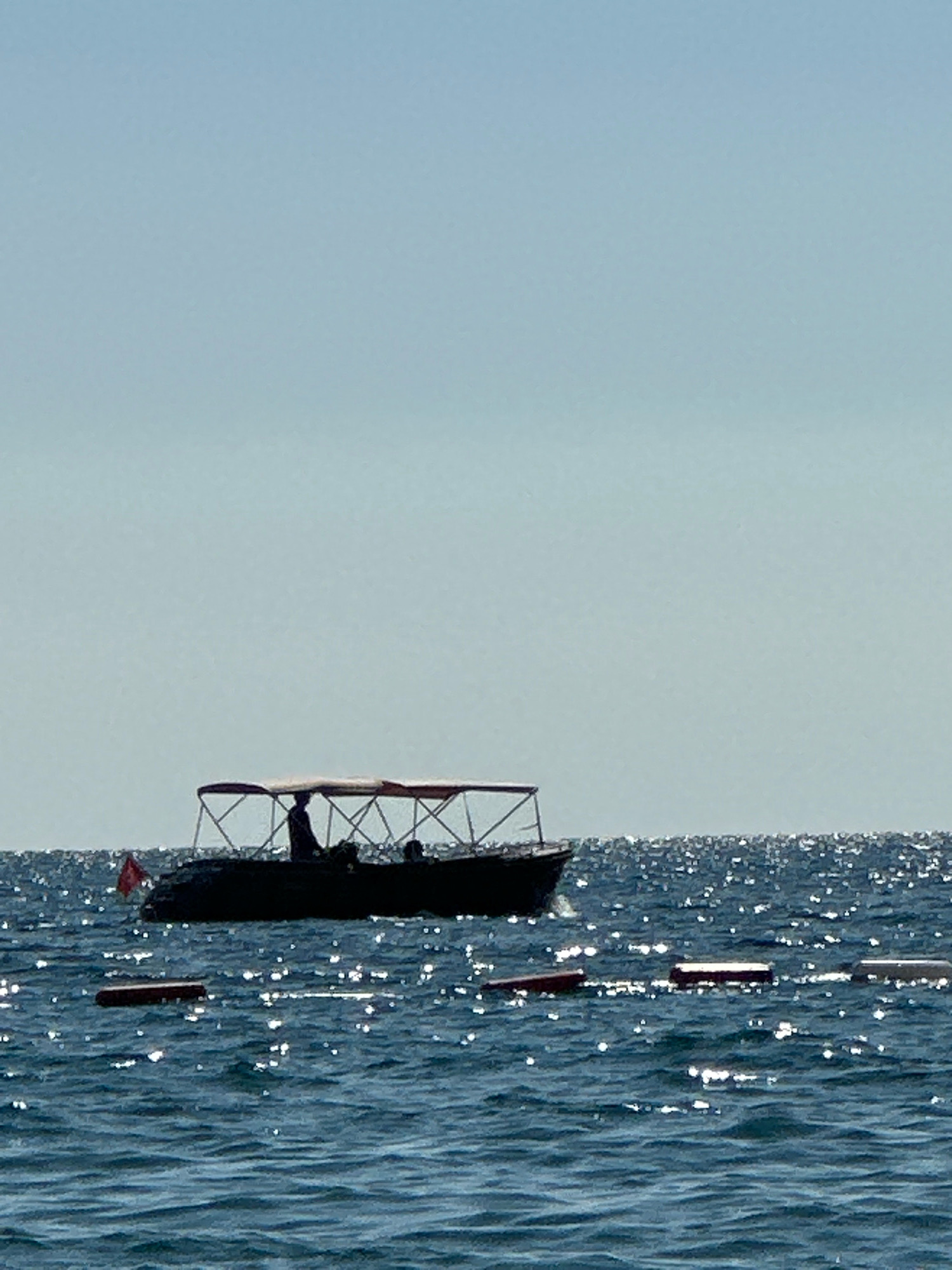 A dark-hulled boat with a canvas canopy is anchored in the bright blue water of Boreti, Montenegro. A single person is visible in silhouette on the boat, near the front. A small red flag is attached to the port side of the boat. The water is choppy with many points of light reflecting off the surface. In the foreground, several buoys or markers are floating in the water, appearing as horizontal red and white rectangular shapes. The sky is a clear, pale blue.