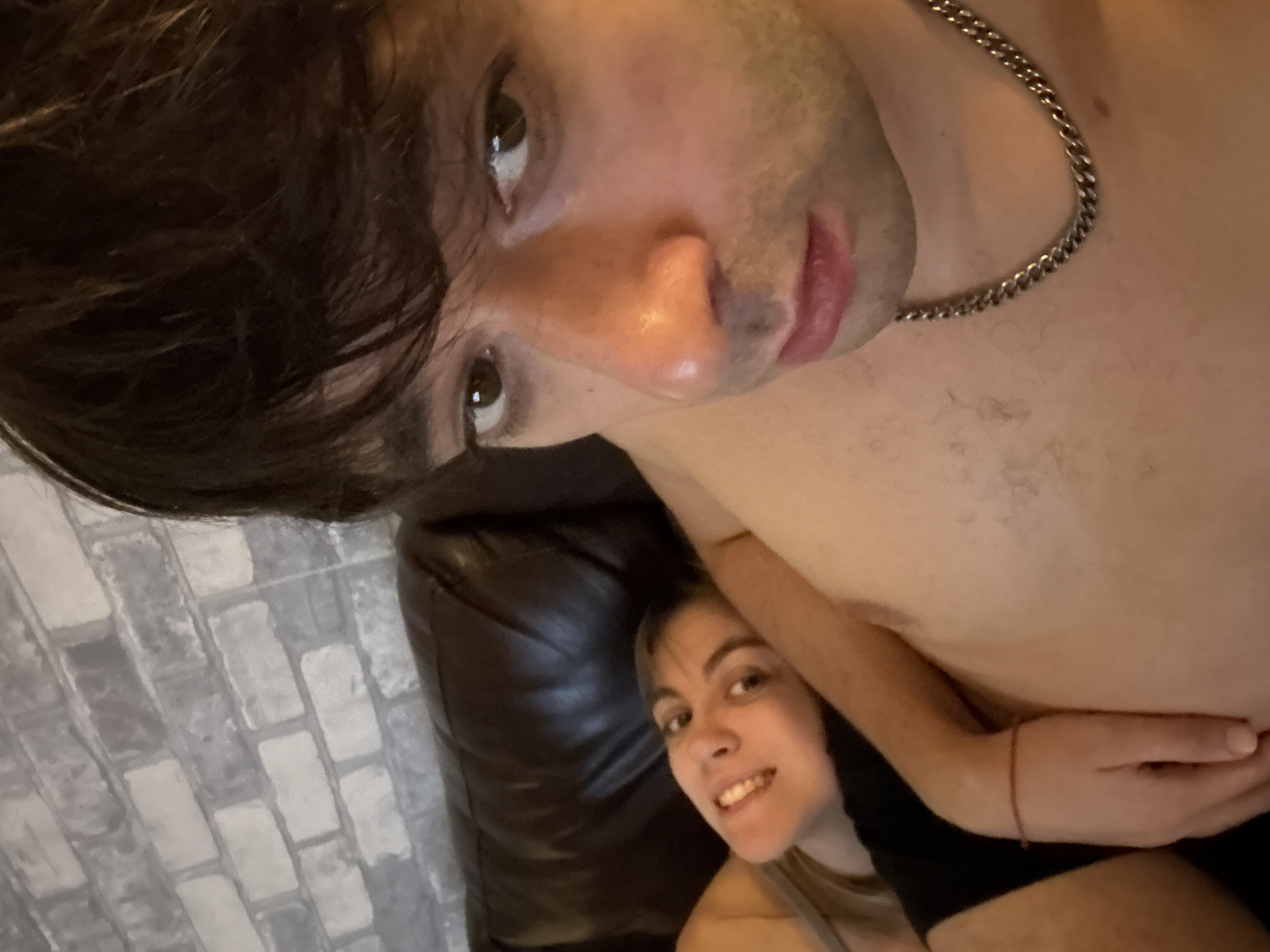 Two individuals are positioned on a brown leather couch, with a brick-patterned wall in the background. The person on the right is a male with dark hair, looking towards the camera with a neutral expression. They are wearing a silver chain necklace. The person on the left is a female with blonde hair, also looking towards the camera, with a slight smile. The scene appears to be indoors. There is no indication of the city of Moscow, Russia.
