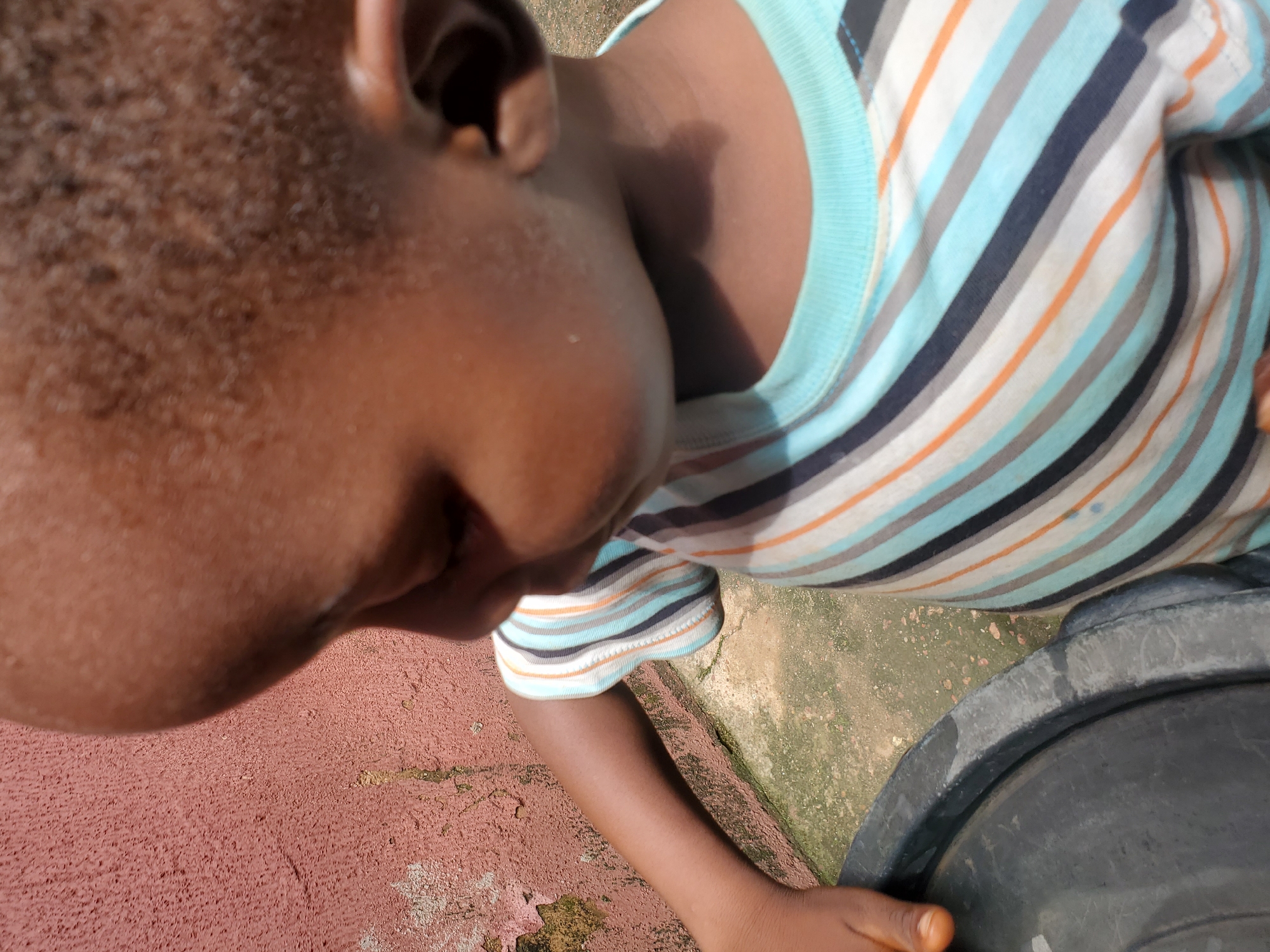 A close-up, high-angle shot captures a young child's head and torso, positioned with their face turned downwards and slightly to the right. The child is wearing a horizontally striped t-shirt in shades of light blue, grey, dark blue, and orange. Their left ear is visible, and part of their neck and shoulder are in focus. The child's skin tone is dark.
The background features textured, reddish-pink ground on the left, likely concrete or pavement, with some patches of moss or dirt. To the right, there's a large, dark, round object, possibly a plastic container or bucket, with a textured rim. The ground here appears to be a mix of concrete and discolored, possibly damp, earth with green moss. The lighting suggests it is daytime, with a strong light source casting shadows. No text is visible in the image. The overall scene gives the impression of an outdoor setting, possibly a yard or play area in Jos, Nigeria. The child's posture and the close-up perspective could suggest a moment of rest or quiet observation.
