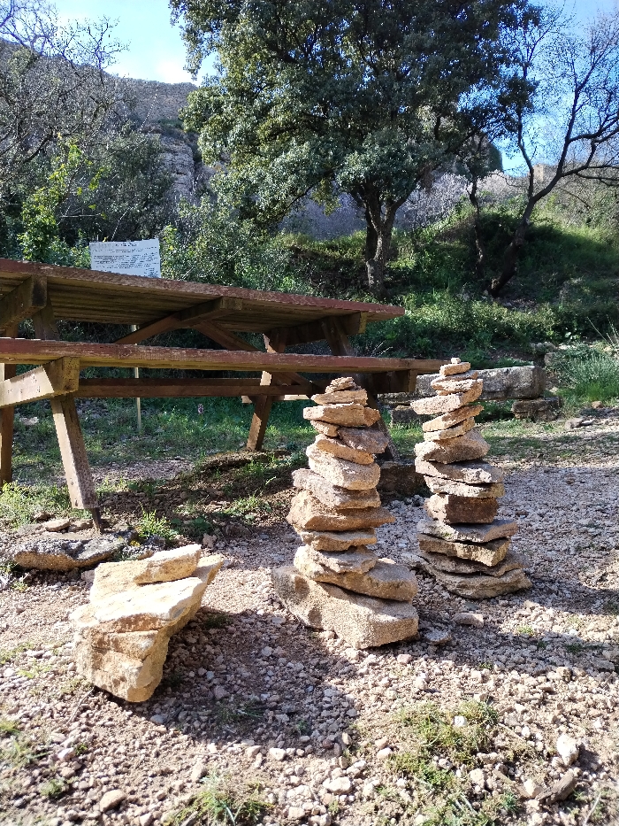 This image captures an outdoor scene, likely a picnic or rest area in a natural setting, under bright daylight. The ground in the foreground and midground is composed of light-colored gravel and small stones, interspersed with patches of green grass.
In the immediate foreground, to the left, is an informal pile of several light brown, irregularly shaped stones. Slightly further back and to the right, are two prominent cairns, or stacks of flat, light brown stones, carefully balanced one on top of the other, forming tall, slender columns. The cairn on the left appears slightly taller and wider than the one on the right.
Dominating the midground is a sturdy wooden picnic table with attached benches, made of thick, weathered planks. Its construction features diagonal supports beneath the tabletop and benches. The table is oriented diagonally to the left, with its length extending towards the left side of the frame.
Behind the picnic table, to the left, a white sign is visible. It appears to have black text or symbols on it, but the details are too small and blurred to be legible. The sign is mounted on a post or frame, slightly obscured by surrounding foliage.
The background consists of dense vegetation and hilly terrain. A variety of trees with green leaves and some with bare branches are visible, suggesting a mix of deciduous and evergreen species. The foliage is lush and green, indicating a healthy natural environment. Behind the trees and vegetation, a rocky or mountainous slope rises, covered in sparse greenery. The sky above is a clear, bright blue, suggesting good weather conditions. Sunlight is strong, creating distinct shadows on the ground and highlighting the textures of the stones and wood. The overall impression is one of a serene and natural outdoor space, possibly a trailside stop or a designated recreation area.