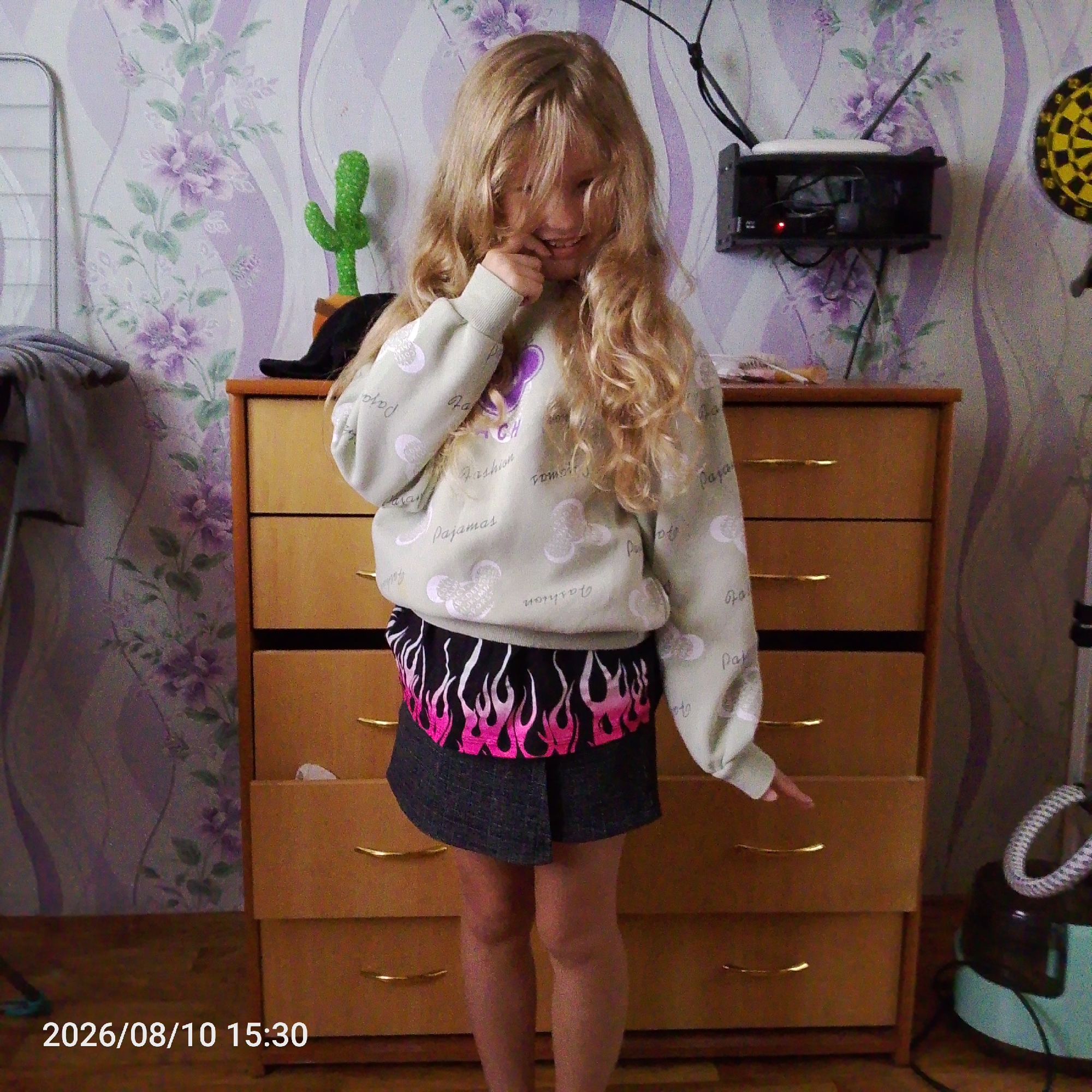 A young girl with long, wavy blonde hair stands in front of a wooden dresser. She is wearing a light green sweatshirt with a pattern of hearts and text, and a black skirt with pink flames. She is smiling and appears to be touching her mouth. Behind her, the wall is decorated with purple floral wallpaper. To the left of the girl, a green cactus decoration is visible, and to the right, a dartboard is mounted on the wall, as well as a router. A timestamp in the lower left corner reads "2026/08/10 15:30". The scene appears to be indoors, possibly in a bedroom or play area.