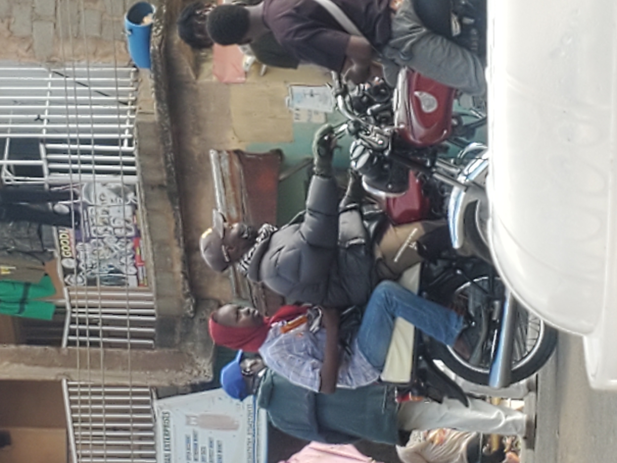The image captures a street scene, likely in Gwong, Nigeria, with several individuals and a motorcycle. In the foreground, a motorcycle is partially visible, with its red fuel tank and chrome accents. A person is seated on the motorcycle, wearing a plaid shirt, blue jeans, and a red headscarf, holding a dark bag. Another person, wearing a grey jacket, scarf, and cap, is also seated on the motorcycle, appearing to be the driver. In the background, a man in a green jacket and blue cap stands beside the motorcycle. Further behind, a building with a metal railing and a blue cylindrical object is visible. The overall environment suggests an urban setting with modest infrastructure.