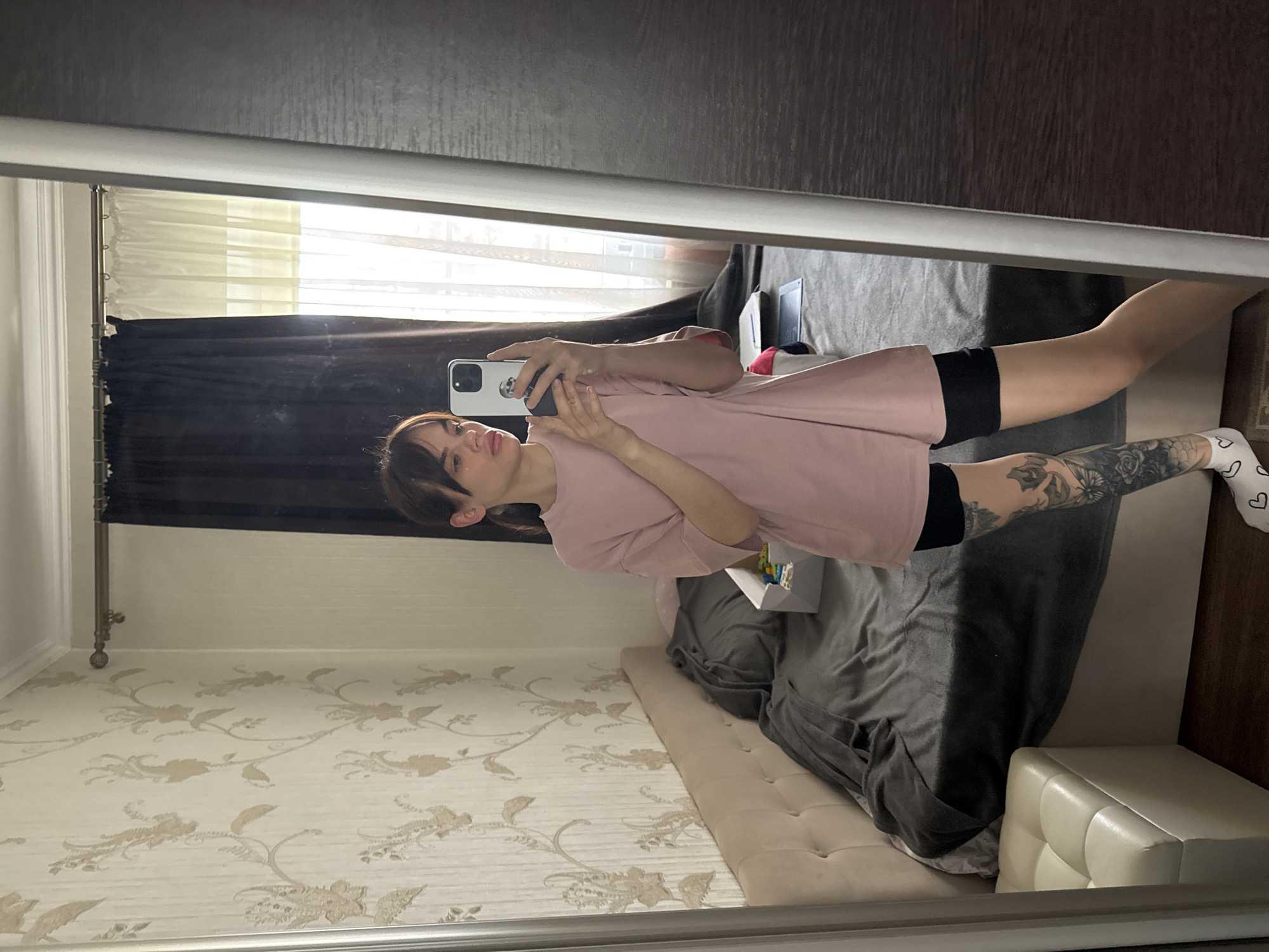 A selfie of a young woman is reflected in a mirror. She is holding a smartphone in her hands, capturing her image. The woman has dark hair with bangs and is wearing a light pink t-shirt and black shorts. Tattoos are visible on her leg. The reflection in the mirror shows a bedroom setting. A bed with a grey duvet is behind her. The wallpaper is patterned with floral designs. A window with curtains is also visible in the background. Rostov-on-Don, Russia cannot be verified as the location based on the image content alone.