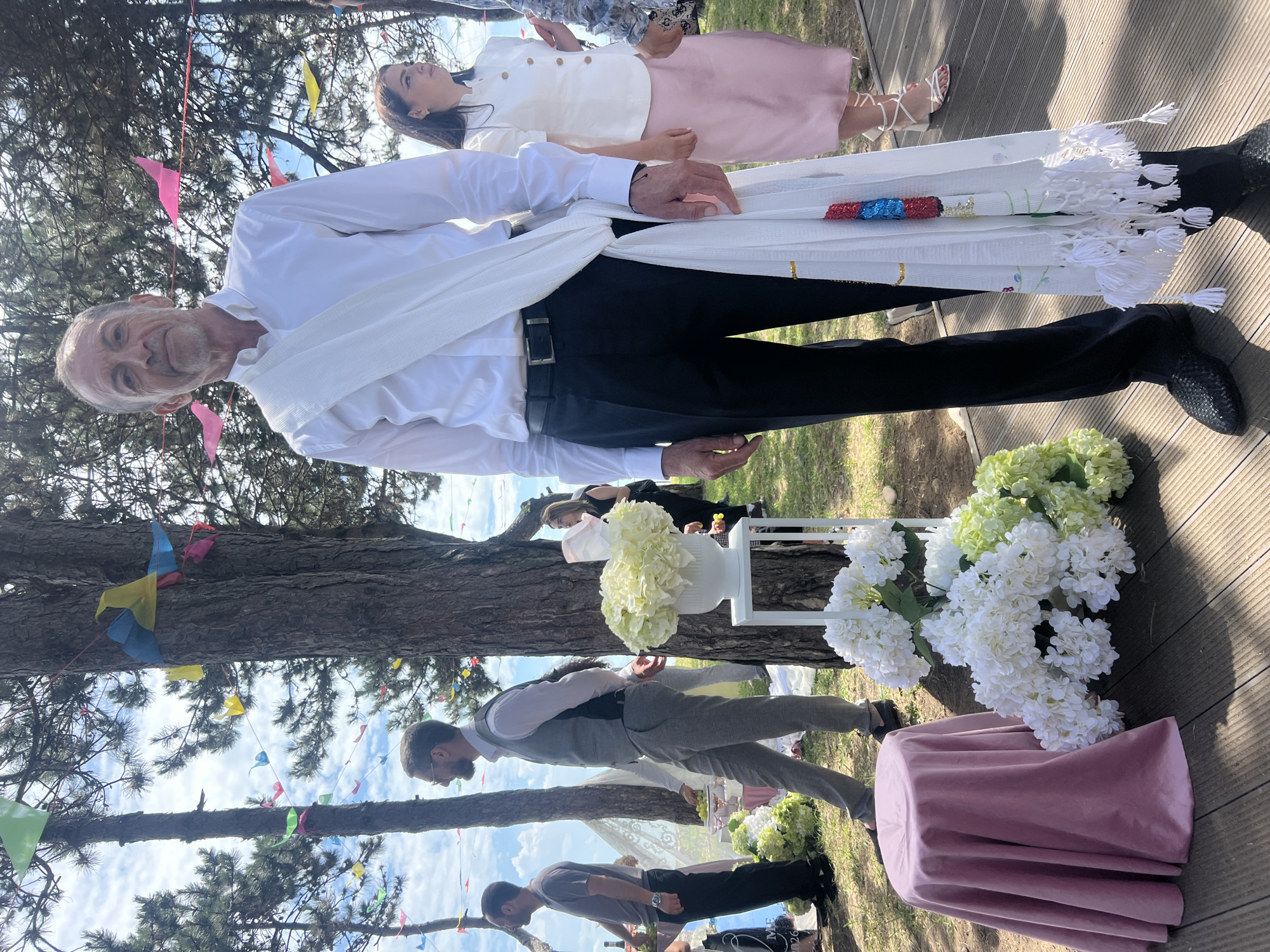 A man in a white shirt and black pants stands on a wooden platform outdoors, holding a decorated white cloth with tassels. He is wearing a white sash draped over his shoulder. Behind him, a woman in a white top and pink skirt stands looking to the side. In the foreground, a pink-covered round table and a white vase filled with white flowers are visible. Trees with colorful triangular flags strung between them are in the background, under a partly cloudy sky. Other people are present further in the background, some standing near tables with food and drinks.