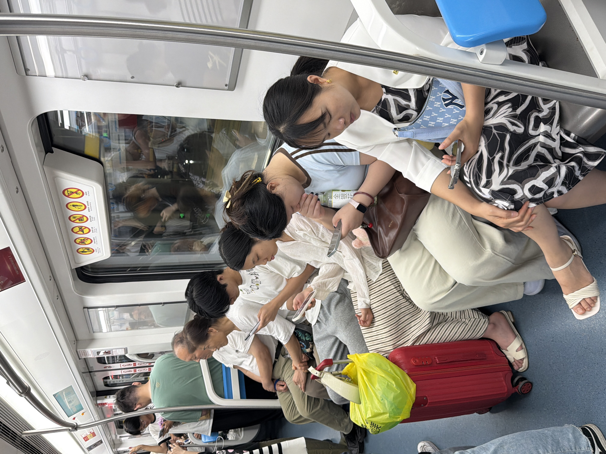 The image depicts the interior of a subway car in Unknown, Unknown, crowded with passengers. The train features white walls and ceiling, a blue floor, and large windows with visible reflections of additional passengers and external elements. Silver handrails and poles are present for standing passengers. Overhead, a panel displays yellow and red circular warning signs depicting actions such as no eating, no leaning, and no littering.
In the foreground, a woman with dark hair tied back sits on the right, wearing a white blazer over a black and white patterned dress and gold hoop earrings. She is looking down at a silver smartphone held in her hands. A light blue purse with a white "NY" (New York Yankees) logo rests on her lap. To her left, another woman with dark hair in a bun is also looking at a smartphone while sipping from a clear plastic bottle labeled "金银花" (Jīn Yín Huā - honeysuckle) with a straw. She wears a light blue t-shirt with brown stripes on the sleeves, a smartwatch on her left wrist, and a dark brown shoulder bag.
Adjacent to them, a woman in a white long-sleeved shirt and striped pants sits with her legs crossed, focusing on a smartphone. A red hardshell rolling suitcase with a bright yellow plastic bag and a white handbag on top is positioned on the floor in front of her. To her left, a young man in a white t-shirt is looking at a smartphone. Further left, an older woman and an older man, both wearing white t-shirts and light brown pants, are seated and looking forward.
On the far left, a young man in a green t-shirt stands facing away, holding onto a silver vertical pole. Behind him, another young man with glasses and a white t-shirt is visible, also looking at a smartphone. Most visible passengers, particularly younger individuals, are absorbed in their smartphones, with minimal direct interaction observed among them.