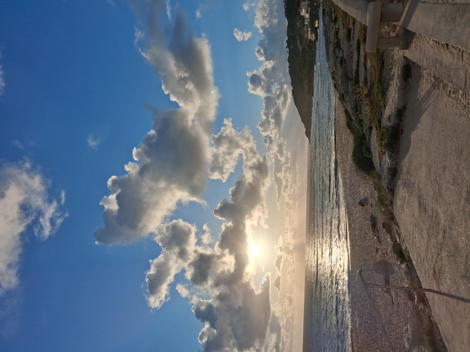 The image depicts a seaside scene, possibly near Figueira da Foz, Portugal, given the coastal environment. A beach with a rocky shore is visible, bordered by a concrete pathway or wall, suggesting a promenade. The sun, low on the horizon, shines through a mix of clouds, creating a bright glare on the water's surface. The sky is a gradient of blue, with the clearest hues at the top and becoming lighter towards the horizon. A mountain or cliff is visible in the background. No people are visible.