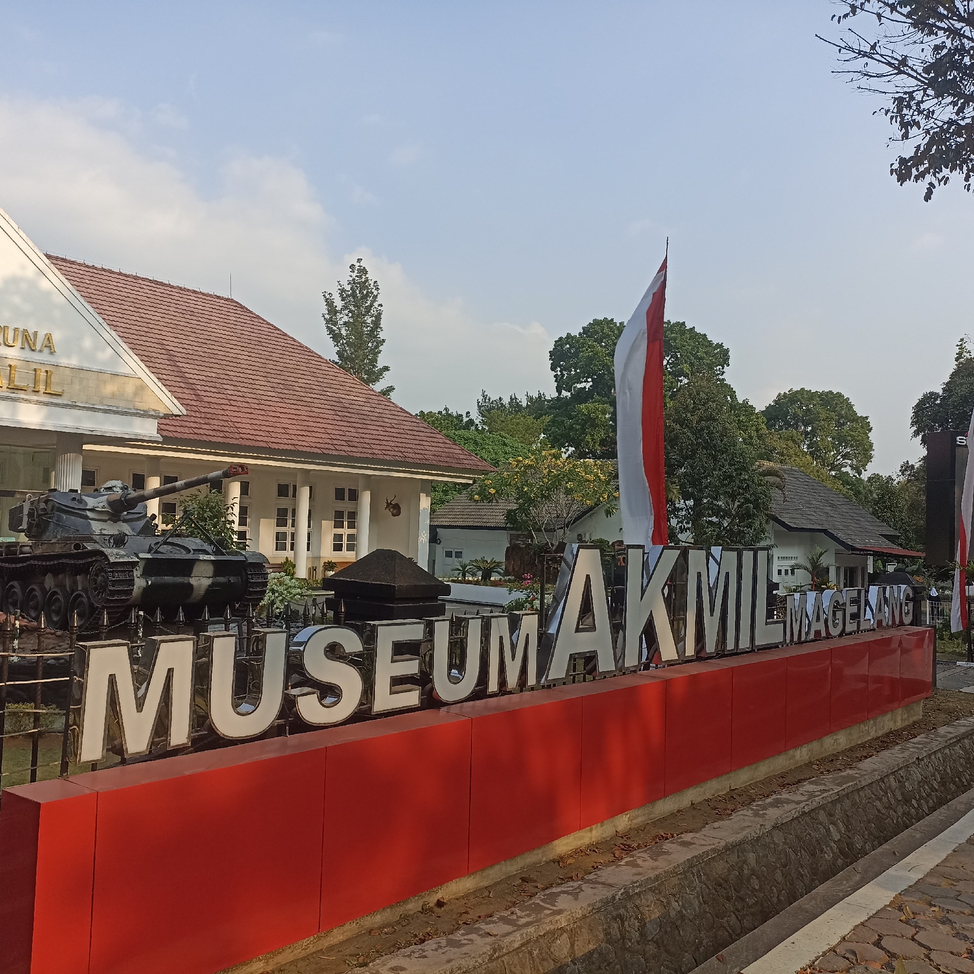 The image shows the exterior of the AKMIL Museum in Magelang, Indonesia, under a bright, partly cloudy sky. In the foreground, a large, raised red base supports prominent white, three-dimensional letters spelling out "MUSEUM AKMIL MAGELANG". Behind this sign, an old military tank is displayed as an exhibit, camouflaged with dark and light green patterns. The tank is positioned in front of a white building with a red tiled roof and white pillars. To the right of the tank, a tall Indonesian flag, red and white, waves on a flagpole. Further in the background, more trees and smaller buildings are visible. The environment appears to be well-maintained, with a paved walkway and a stone-lined drainage ditch in the immediate foreground. No people are clearly visible in the frame.