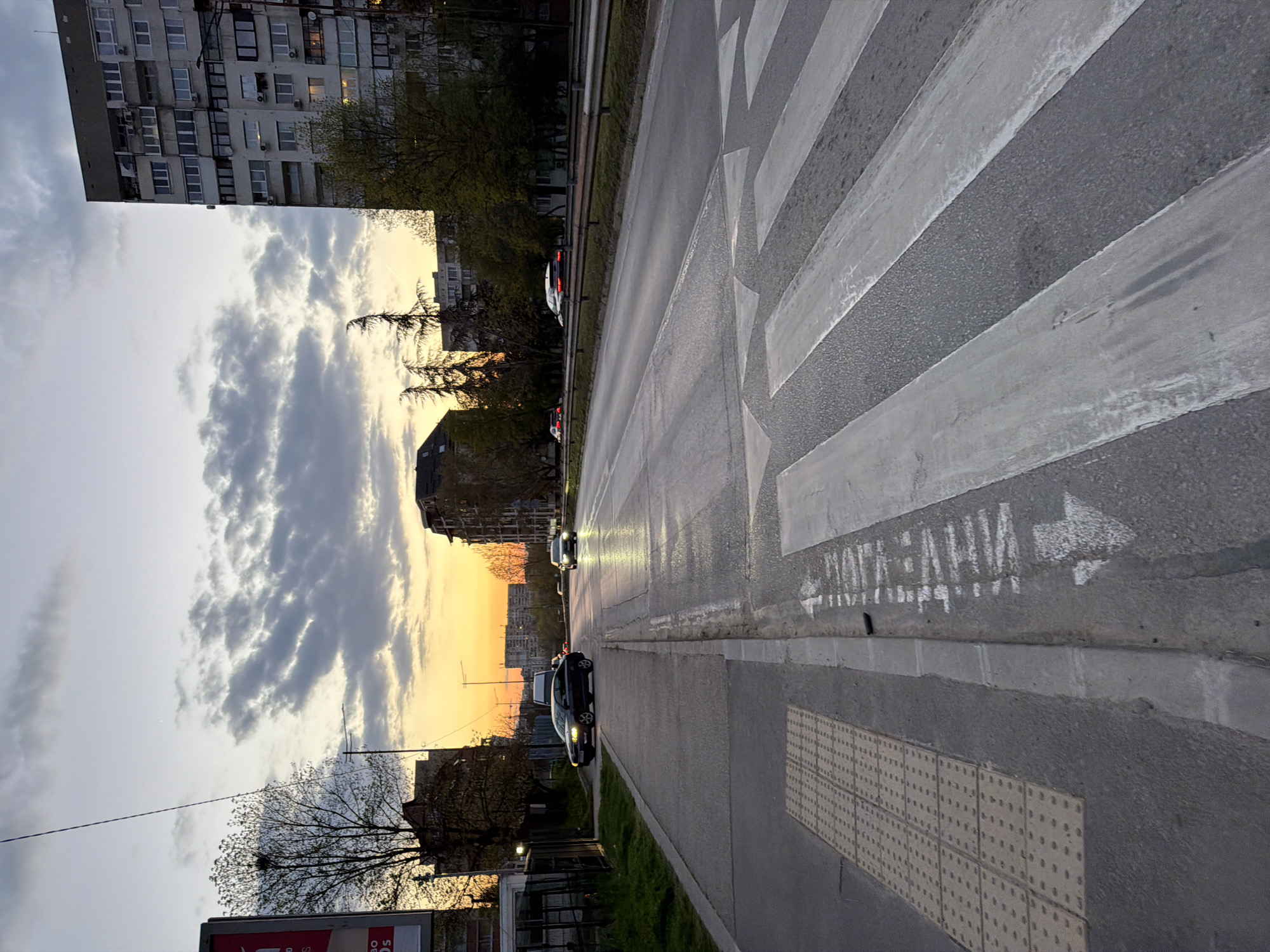 The image captures an urban street scene at dusk or early evening in Sofia, Bulgaria. The sky transitions from a warm, orange-yellow sunset glow on the horizon to an overcast, grey sky overhead, with visible clouds. The street runs diagonally from the lower right towards the upper left, receding into the distance.
In the foreground, a prominent zebra crossing (crosswalk) spans the width of the road, featuring alternating white and grey stripes. Adjacent to the crosswalk, on the sidewalk, is tactile paving designed for visually impaired pedestrians. White Cyrillic text is painted on the asphalt surface next to the crosswalk, which reads "ВНИМАНИЕ ЗАНИ" followed by a white arrow pointing towards the left.
The road is wet, reflecting the car headlights and the sky, suggesting recent rain or damp conditions. Several cars are visible on the road, with at least two in the mid-ground having their headlights on, illuminating the wet asphalt. To the left of the road, a multi-story apartment building with numerous windows and balconies dominates the upper part of the frame, characteristic of Eastern European architecture. Trees, some still bare and others with early spring foliage, line the street and are silhouetted against the sky. Other buildings and power lines are visible further down the street on both sides. The overall mood is quiet and atmospheric, typical of a city transitioning from day to night.