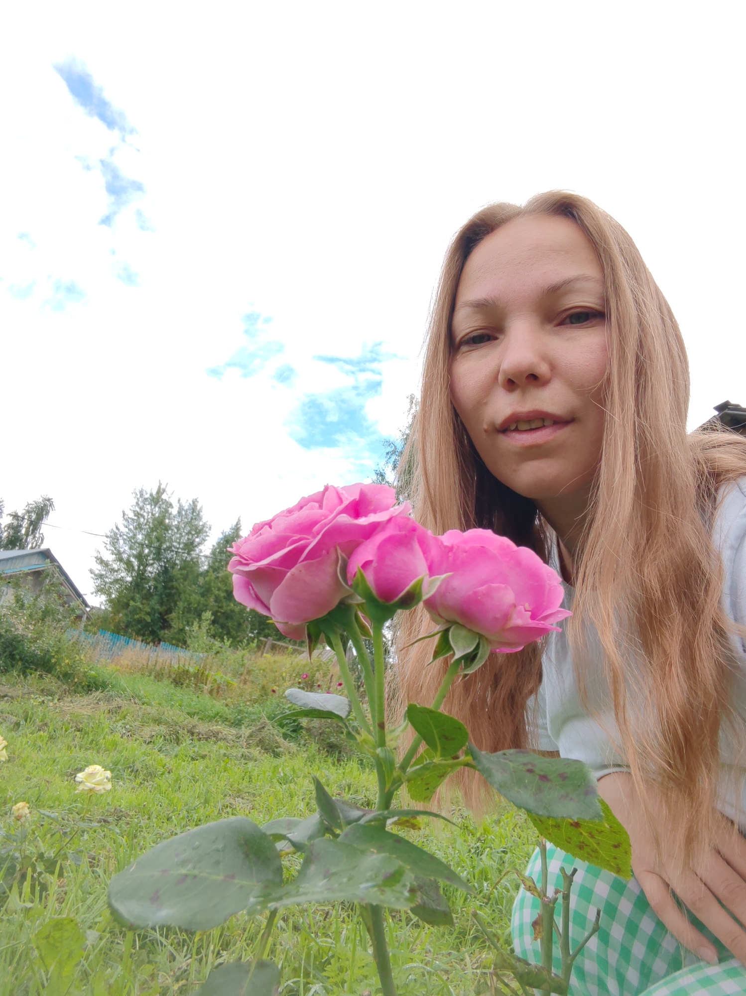 In an outdoor, grassy scene, a fair-skinned woman with long, blonde hair is captured from the chest up, leaning towards the camera. She wears a light-colored shirt and a green and white checkered garment on her lower half. In the foreground, a plant with two pink roses and green leaves is prominently featured. The background shows a yard with more greenery, a fence, trees, and a simple structure that appears to be a house. The sky above is bright with scattered clouds, suggesting a daytime setting. The overall impression is that of a candid snapshot taken in a rural or suburban environment.