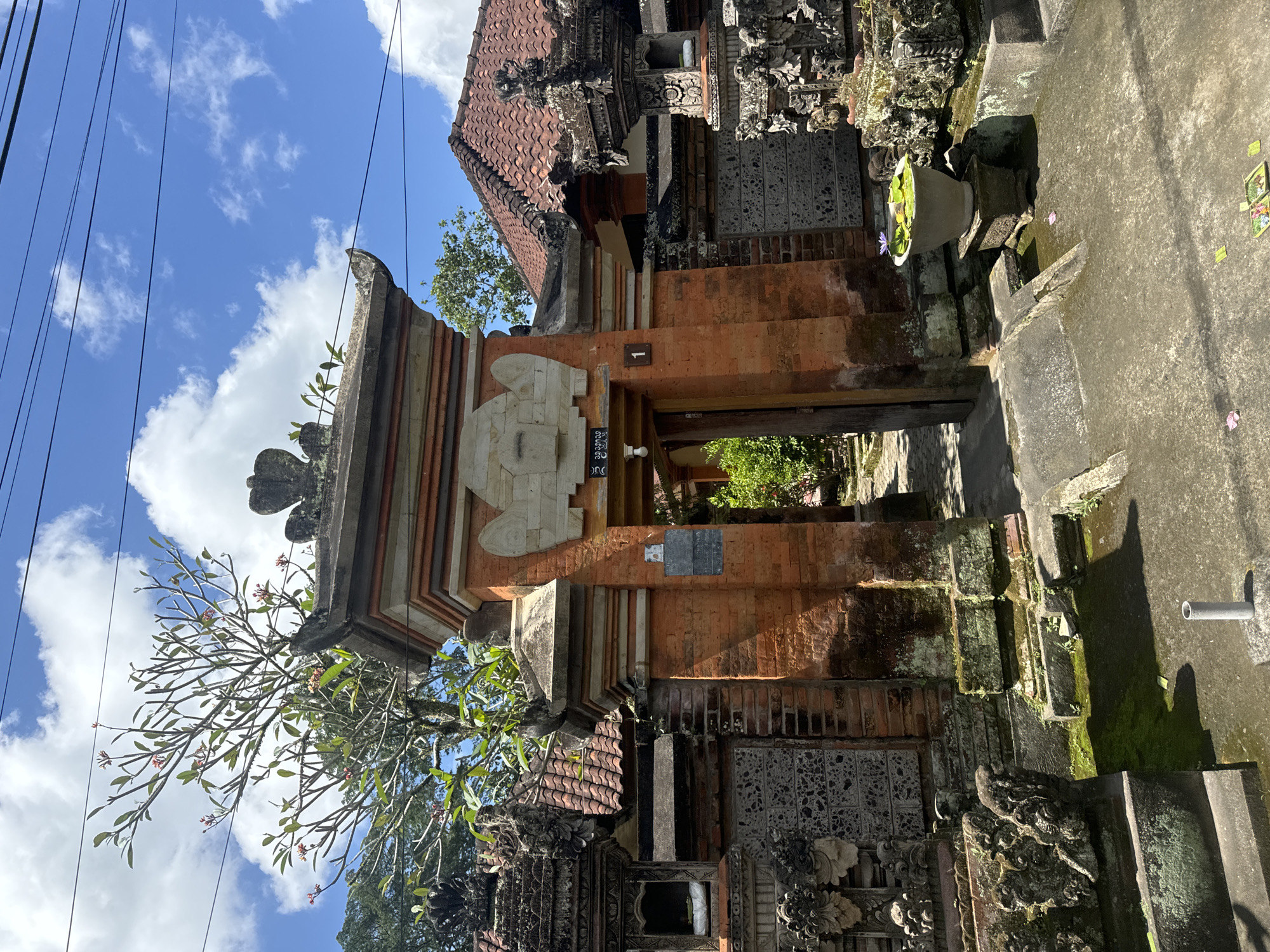 The image showcases a traditional Balinese gate, likely in the Ubud District of Indonesia, characterized by its intricate stone and brickwork construction. The gate is partially shaded, with elements of moss and weathering indicative of age and exposure to the humid climate. Overhead power lines are visible against a backdrop of a blue sky with scattered clouds. A frangipani tree, with its characteristic branches and blossoms, is situated to the left of the gate. The surrounding architecture includes tiled roofs and detailed stone carvings, typical of Balinese design. The atmosphere is suggestive of a historic site or traditional village within the Ubud District.