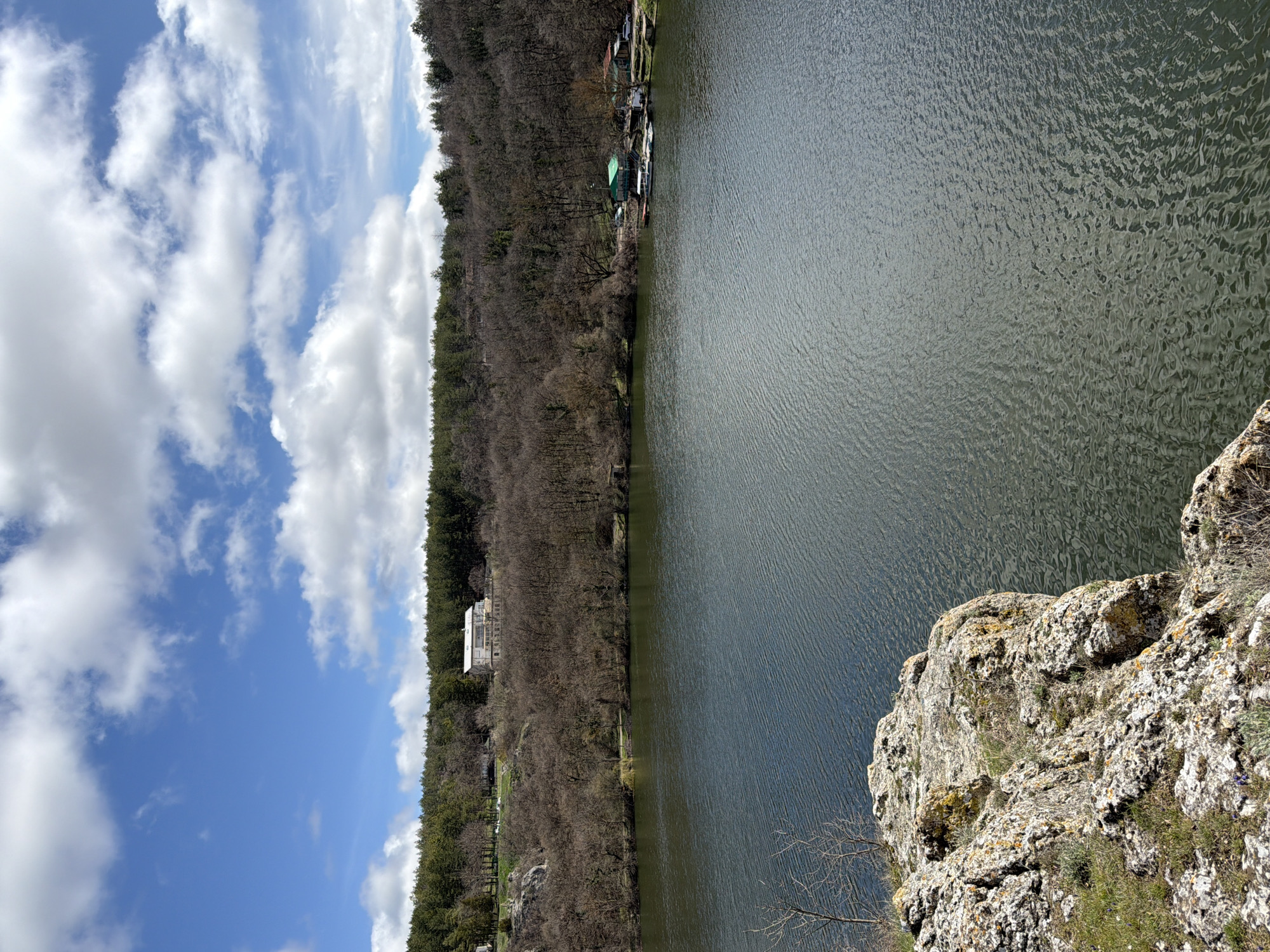 The image displays a panoramic view of a lake or reservoir within Kailuka Park, Pleven, Bulgaria, under a partially cloudy sky. In the lower-left foreground, a rugged, textured rocky outcrop covered with lichen and sparse vegetation occupies a portion of the frame, indicating an elevated vantage point.
The central and lower portions of the image are dominated by the dark green surface of the water, exhibiting numerous ripples. The far bank is densely forested, with a mix of deciduous and coniferous trees. On the left side of the far bank, a prominent two-story white building with a dark roof and visible windows stands on a cleared, grassy area, elevated on a hillside. Below this building, the hillside is covered by bare deciduous trees. Further up the slope, a dense coniferous forest forms the skyline.
On the right side of the far bank, a cluster of smaller, single-story structures with green roofs is visible directly at the water's edge. Several small boats are moored in the water adjacent to these buildings. A white sedan is parked on the bank near the structures. The hillside behind these buildings also features bare deciduous trees, transitioning into a coniferous forest at the higher elevations. The sky above is clear blue with scattered white cumulus clouds. No individuals or active interactions are visible in the scene.